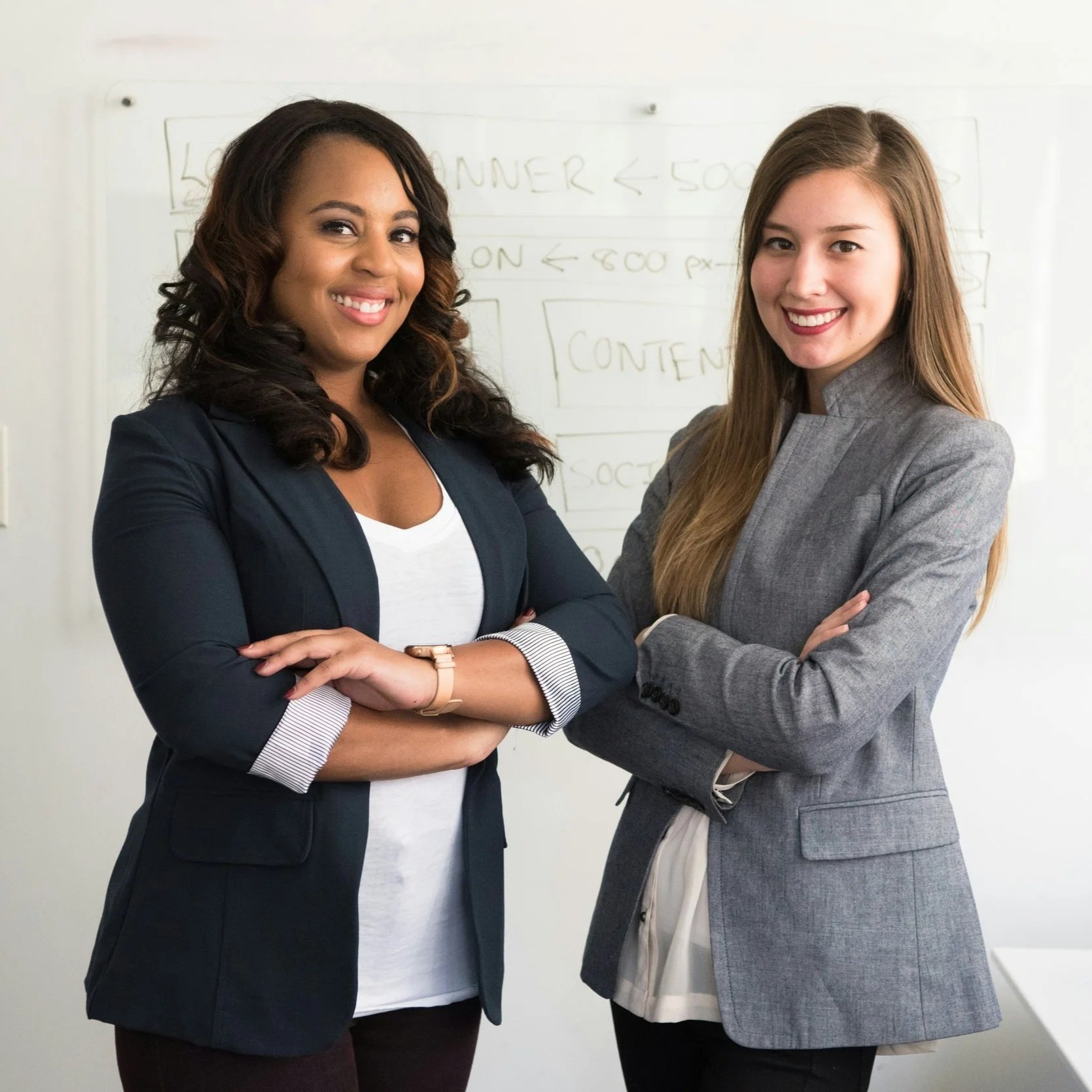 Two women smiling and standing close with arms crossed in front of a whiteboard filled with handwritten notes and diagrams.