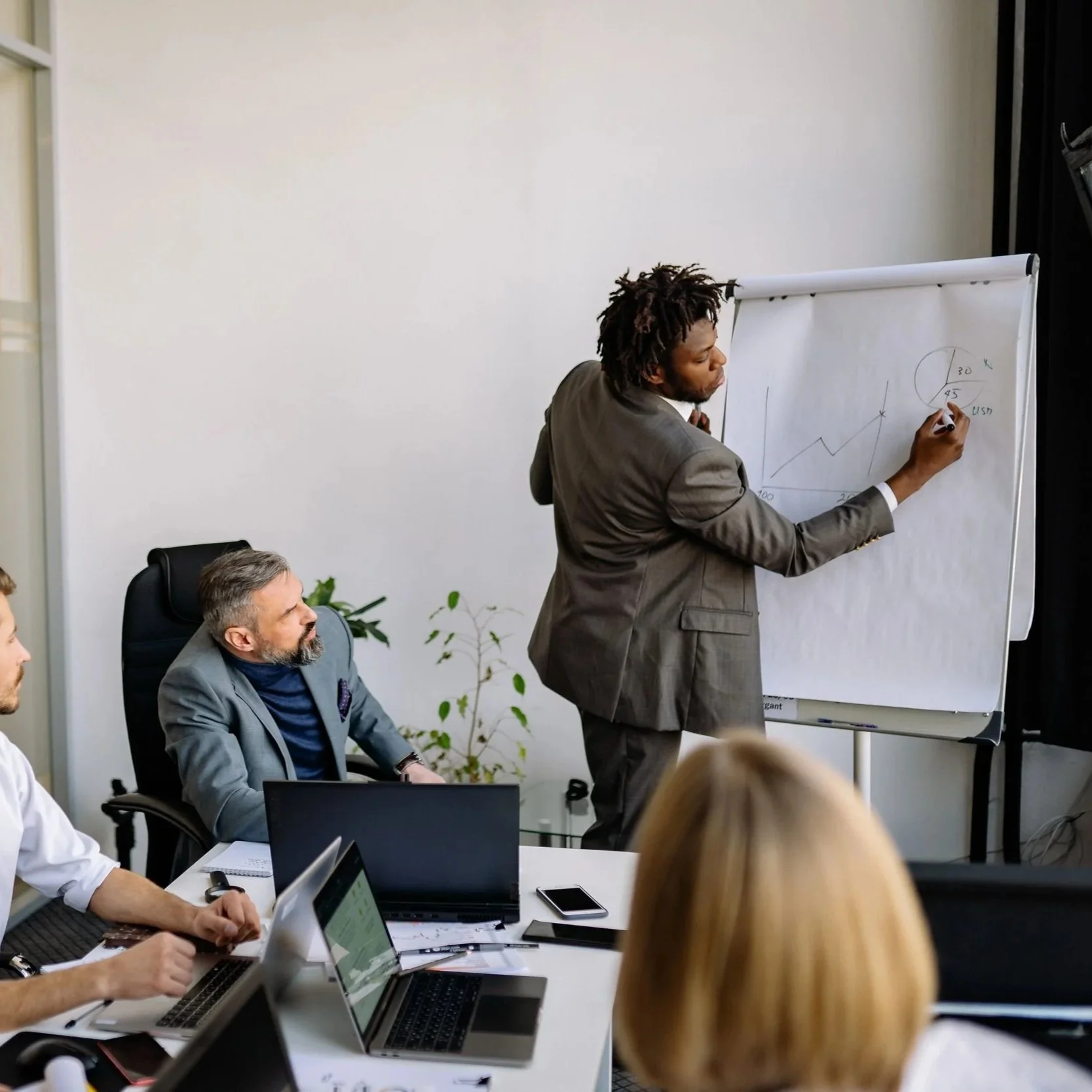 A business meeting in a conference room with a man in a gray suit giving a presentation on a whiteboard while others listen and take notes on laptops and tablets.