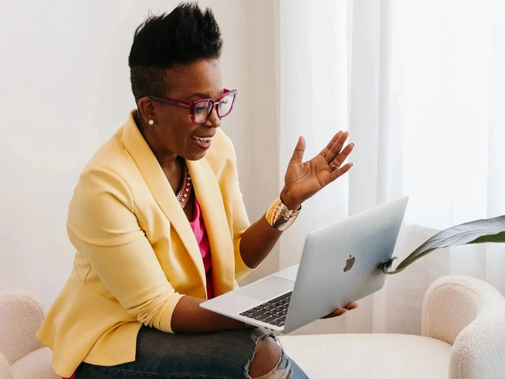 A woman with short black hair and glasses, wearing a yellow blazer, colorful earrings, and ripped jeans, sitting on a white sofa, smiling, during a video call on her open laptop with a plant and white curtains in the background.