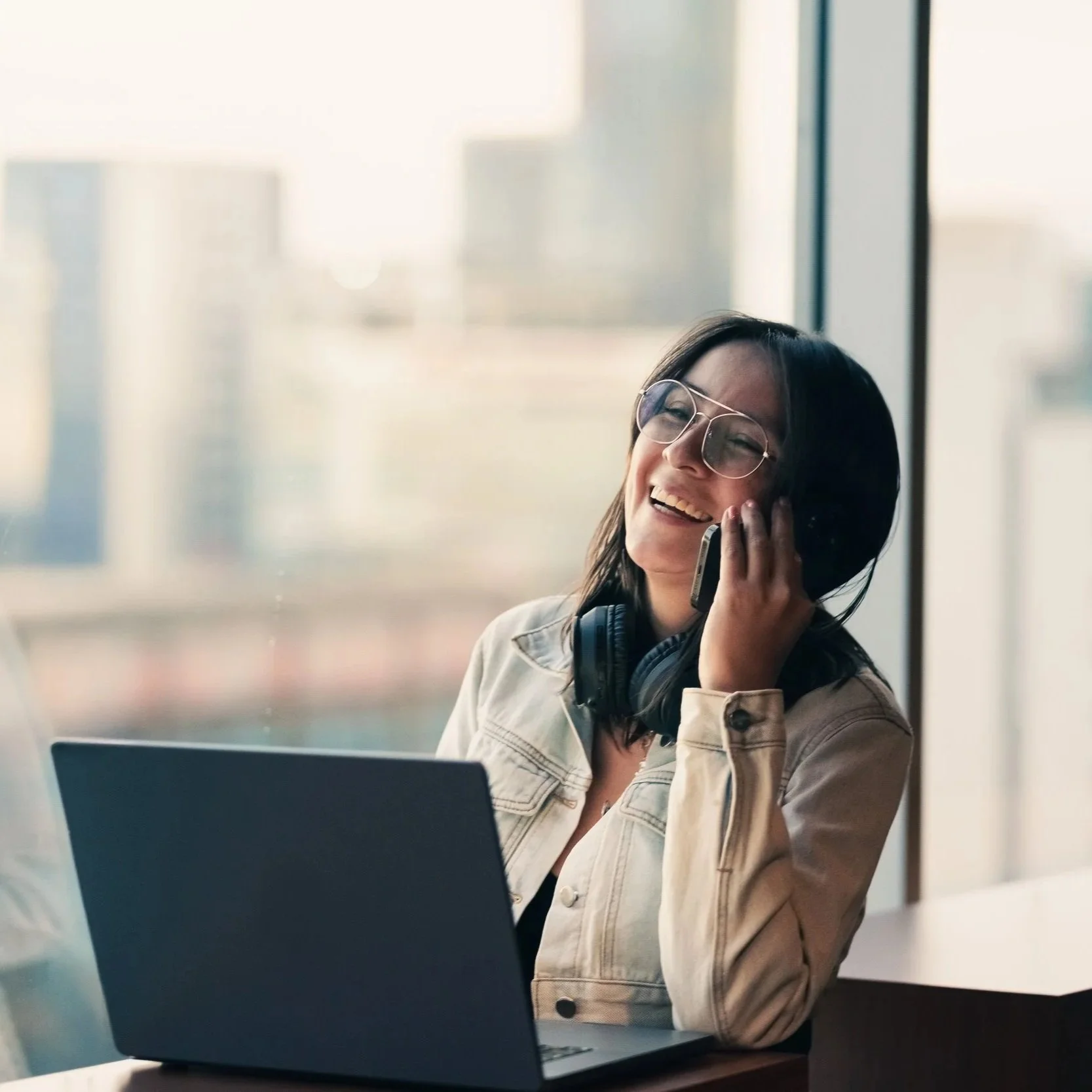A woman with glasses and headphones on her neck, smiling and talking on her phone while working on a laptop in an office with large windows.