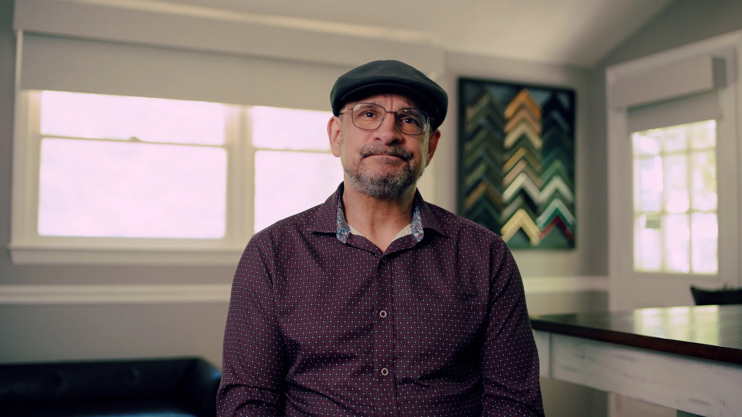 A middle-aged man with glasses, facial hair, and wearing a dark cap and patterned shirt, standing in a well-lit room with large windows, artwork on the wall, and a table in the foreground.