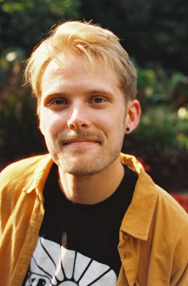 A young man with blonde hair and a beard, smiling, wearing a yellow shirt over a black graphic t-shirt, outdoors with blurred greenery in the background.