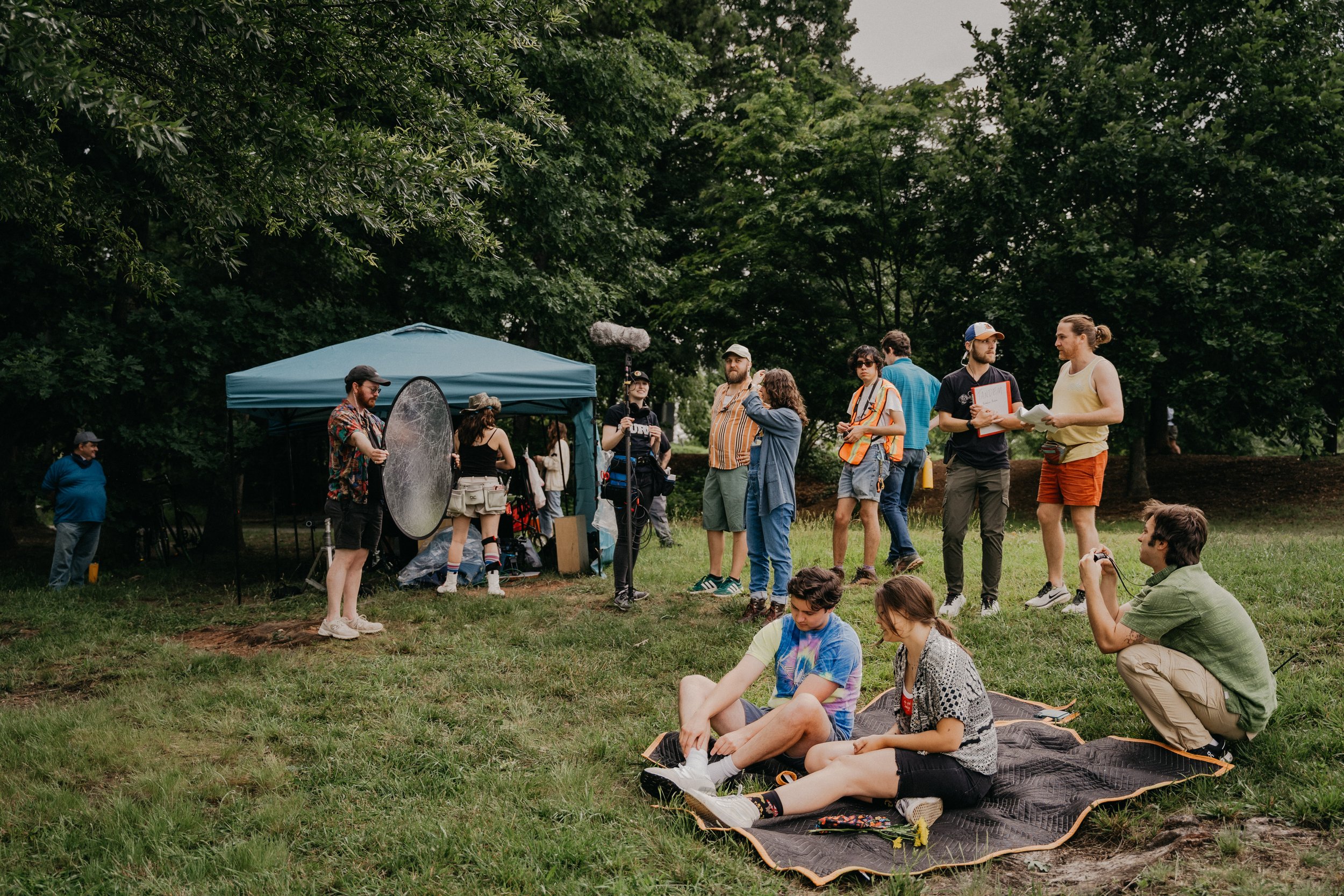 Film crew on a park set filming a scene, with people sitting on a blanket in front, and equipment and crew members gathered under a blue canopy.
