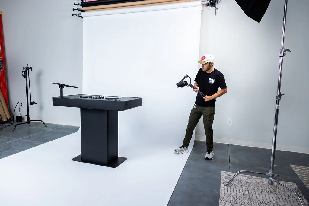 Photographer holding a camera stabilizer in a studio with a white backdrop, a podium, and professional lighting equipment.