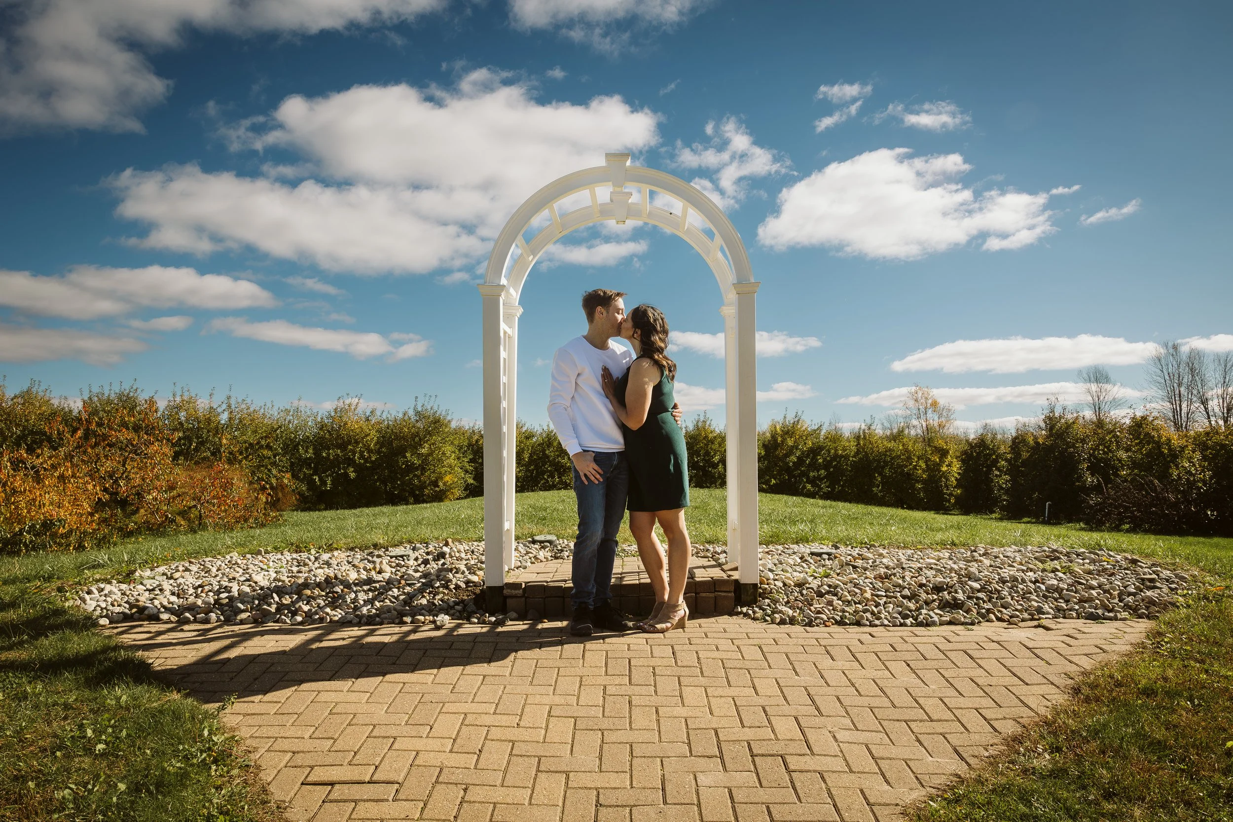 A couple sharing a kiss under a white outdoor arch on a sunny day with blue sky and scattered clouds. Engagement photoshoot.