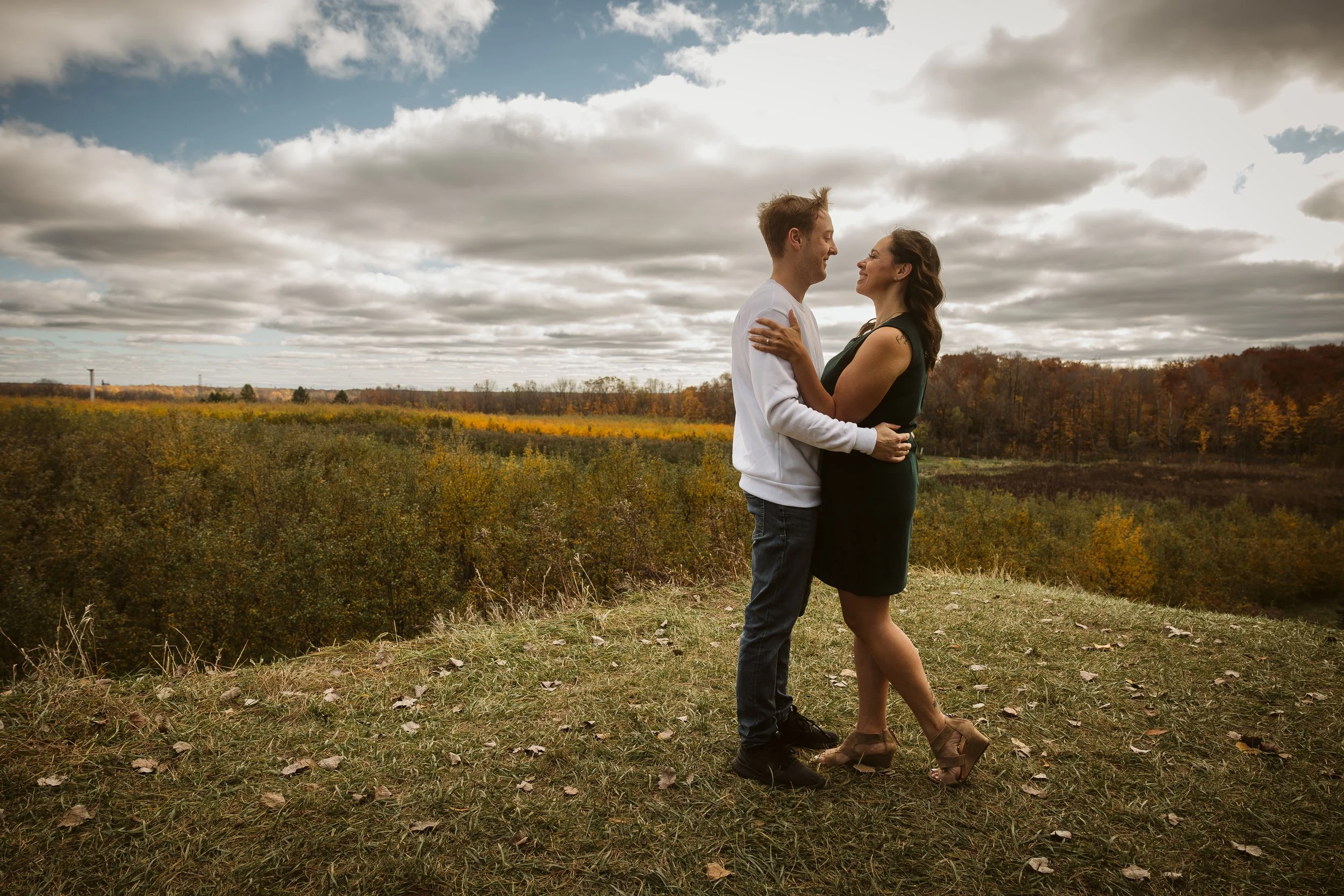 A couple embracing outdoors on a grassy hill with an autumn landscape and cloudy sky in the background. Engagement photoshoot.