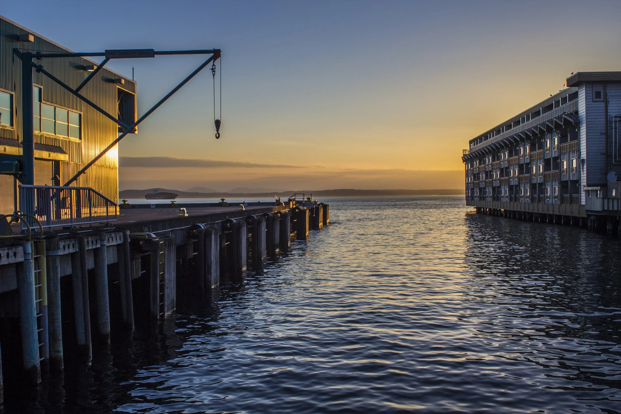 View of a harbor at sunset with a crane on the left, a body of water in the center, and a building on the right, with distant hills on the horizon. Seattle, Washington. Landscape photography.