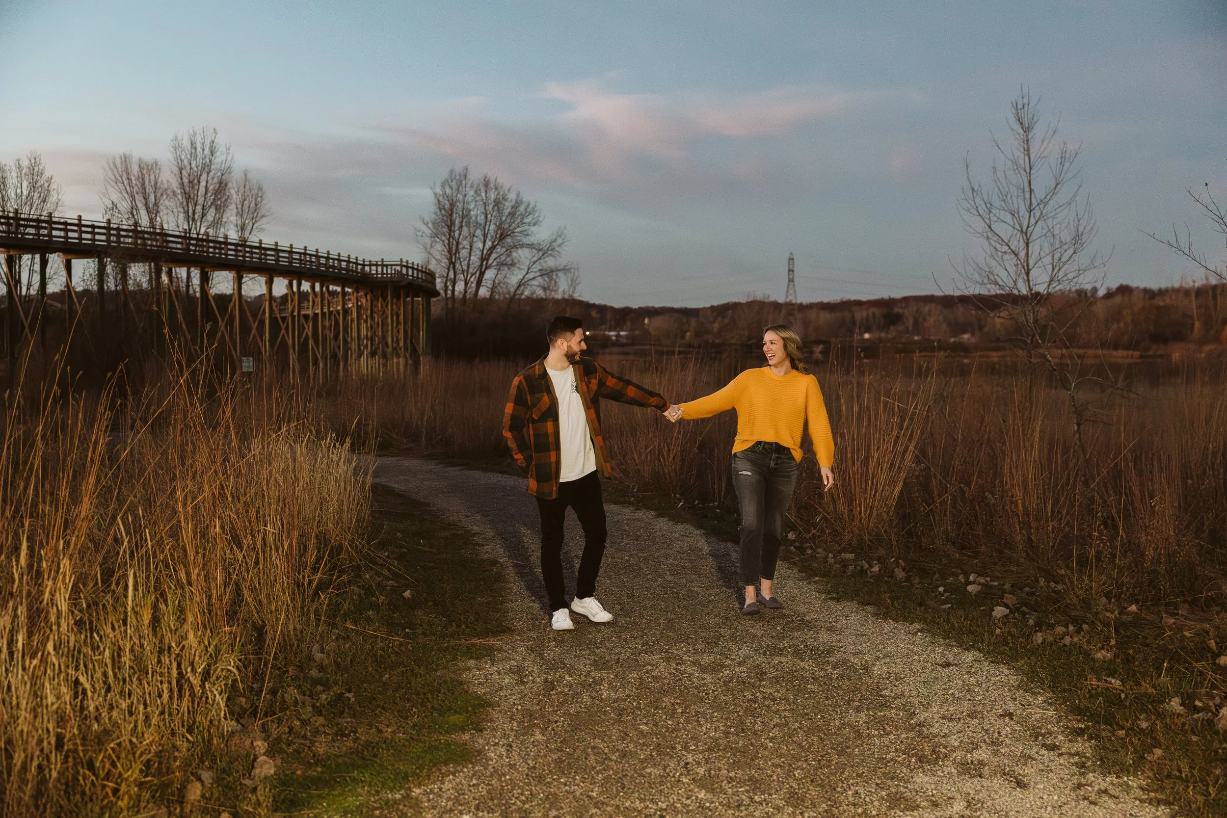 A man and woman walking along a dirt path holding hands, surrounded by tall grass and trees, with a bridge in the background during dusk. Engagement photoshoot.