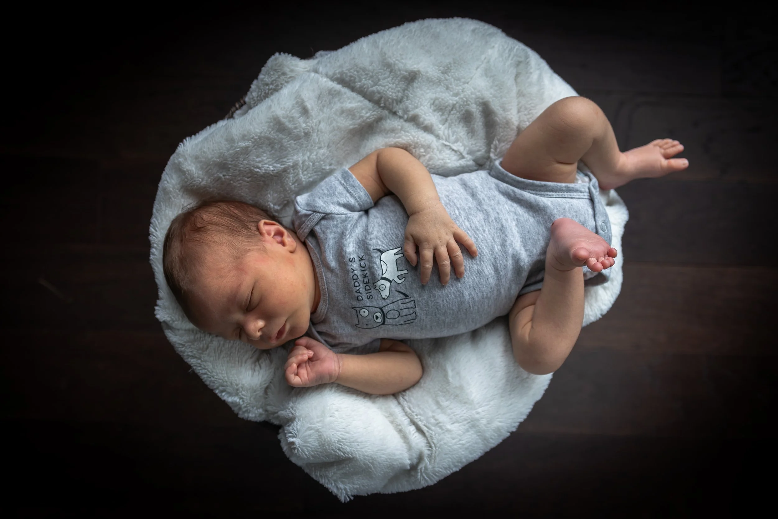 A baby sleeping on a soft, white blanket on a dark wooden floor. Newborn photography. Lansing, Michigan.
