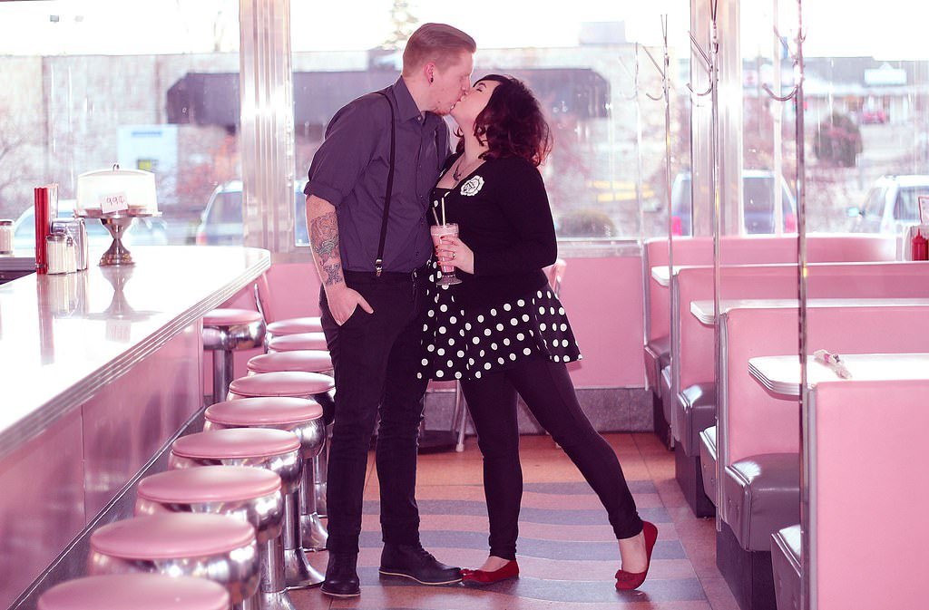 A couple sharing a kiss in a vintage-style diner with pink booths and stools, large windows in the background, and 50s decor. Engagement photoshoot.