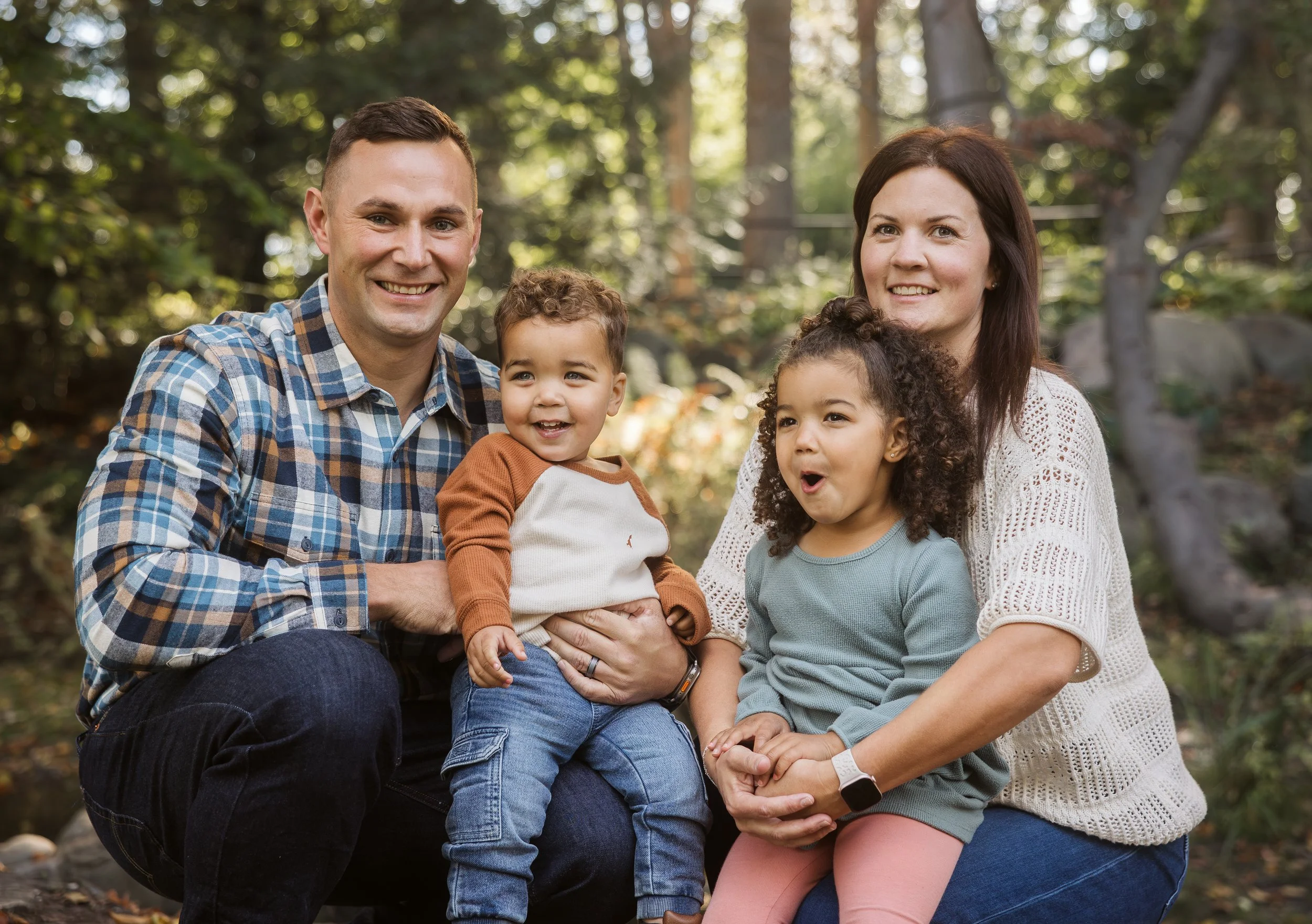 Family of four smiling outdoors in a wooded area, with two adults and two children. Family photography. Grandville, Michigan.