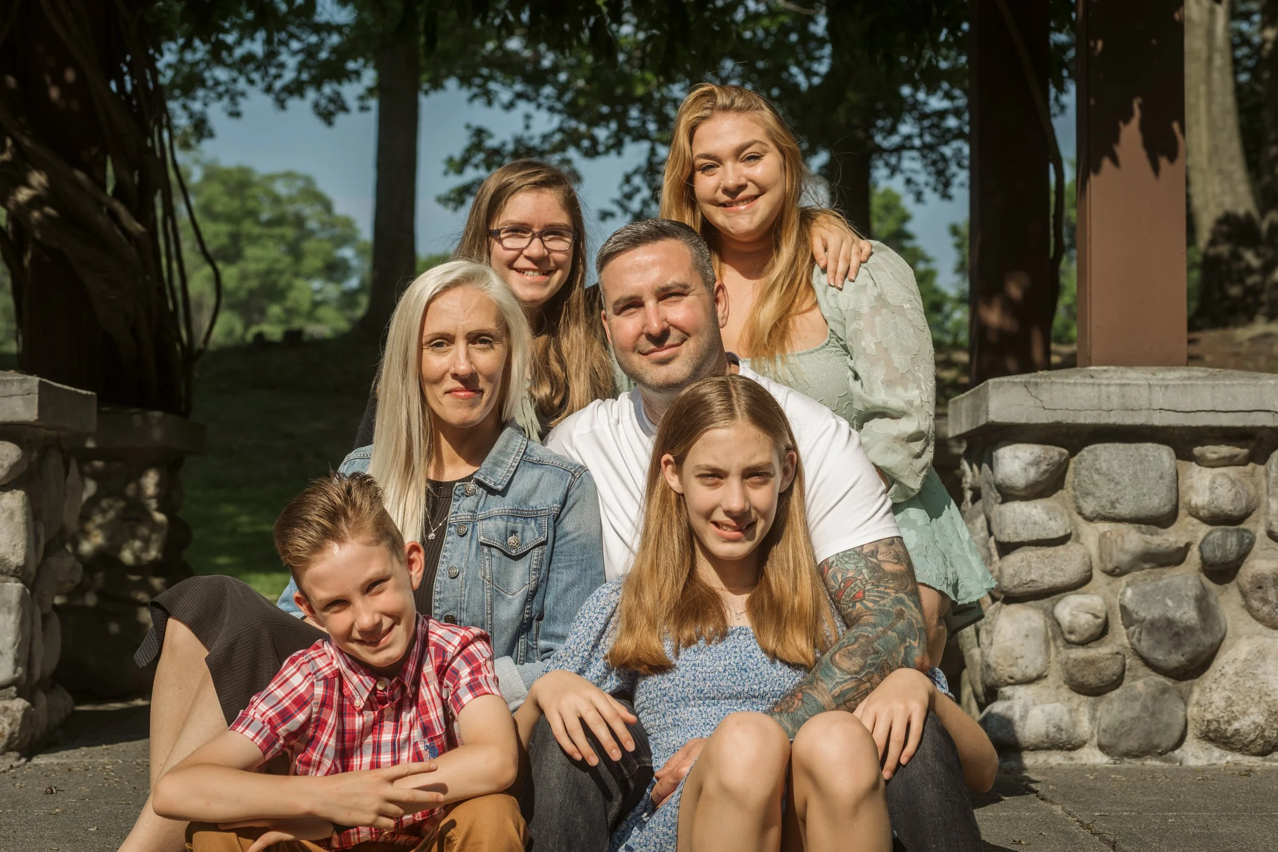 Family group photo outdoors on a sunny day, with two adults and four children sitting and standing on stone steps surrounded by trees. Family photography. Grandville, Michigan.