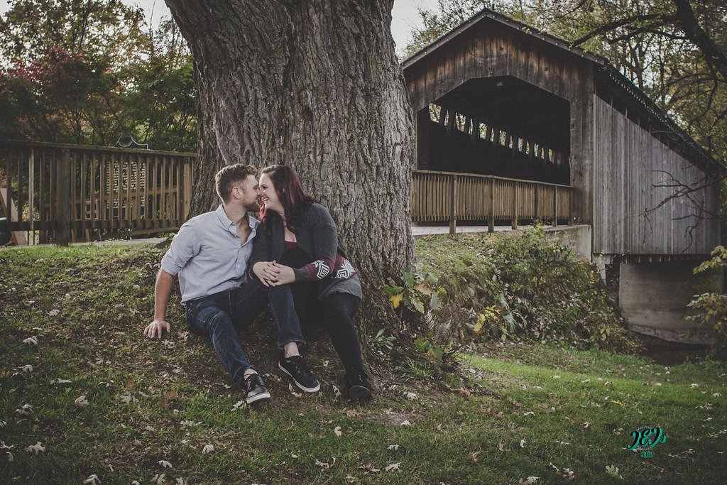 A couple sitting on the grass under a large tree, leaning against its trunk, with a wooden covered bridge in the background. They are sharing a close, affectionate moment and smiling at each other. Engagement photoshoot.