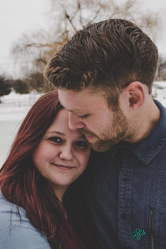 A young couple embracing outdoors, with the woman smiling and the man gently touching his forehead to hers, in a snowy landscape. Engagement photoshoot.