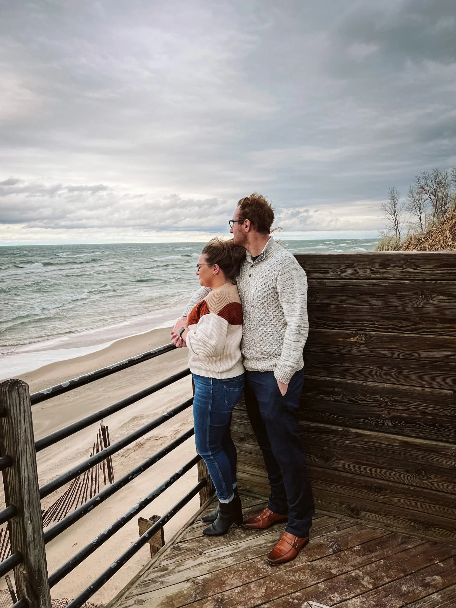 A couple standing on a wooden deck at the beach, looking out at the ocean with clouds overhead. Engagement photoshoot.
