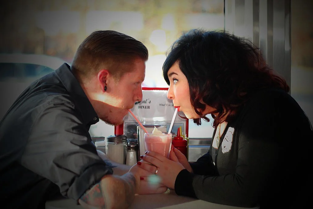 A man and woman sharing a milkshake at a diner, leaning close to each other with straws in the drink. Engagement photoshoot.