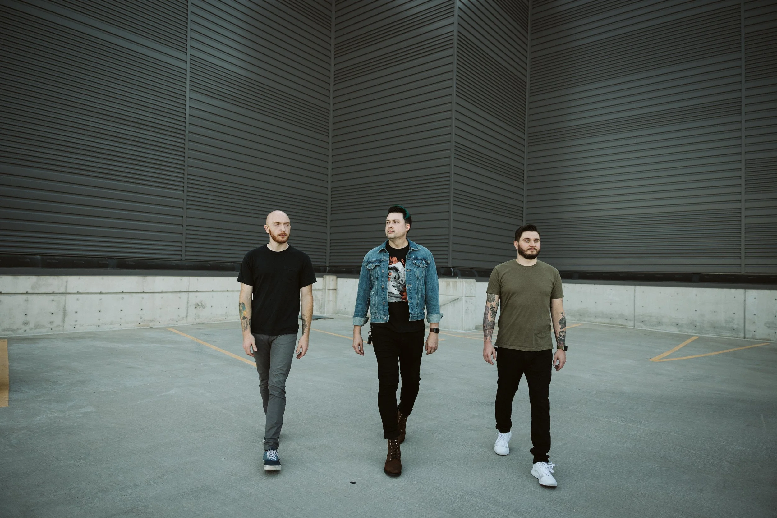 Three men walking in an empty parking lot against a dark, industrial building with horizontal siding. Band photography. Downtown Grand Rapids, Michigan.