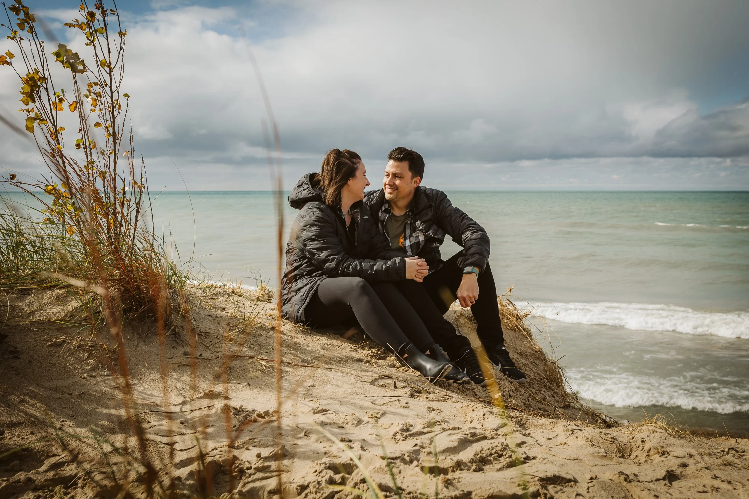 A couple sitting on a sandy beach near the water, smiling and holding hands, wearing dark jackets, with cloudy skies overhead. Couple photography. Lake Michigan.