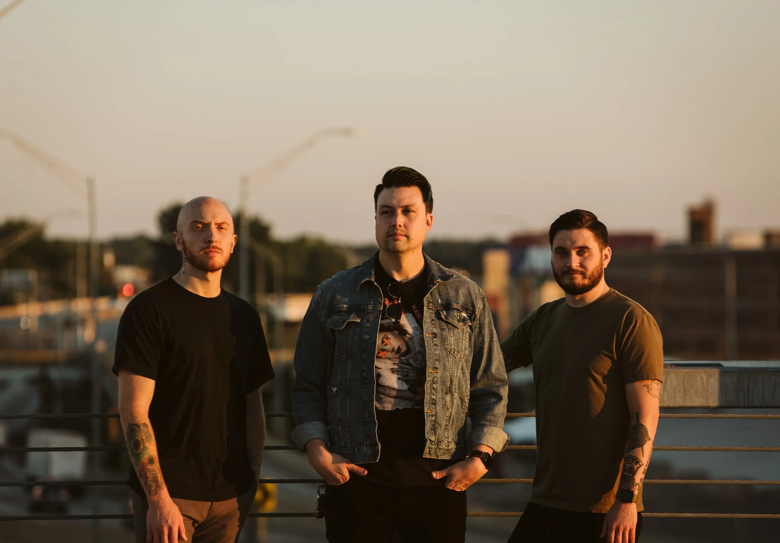 Three men standing outdoors on a rooftop during sunset, facing the camera with serious expressions, with buildings and streetlights blurred in the background. Band photography. Downtown Grand Rapids, Michigan.