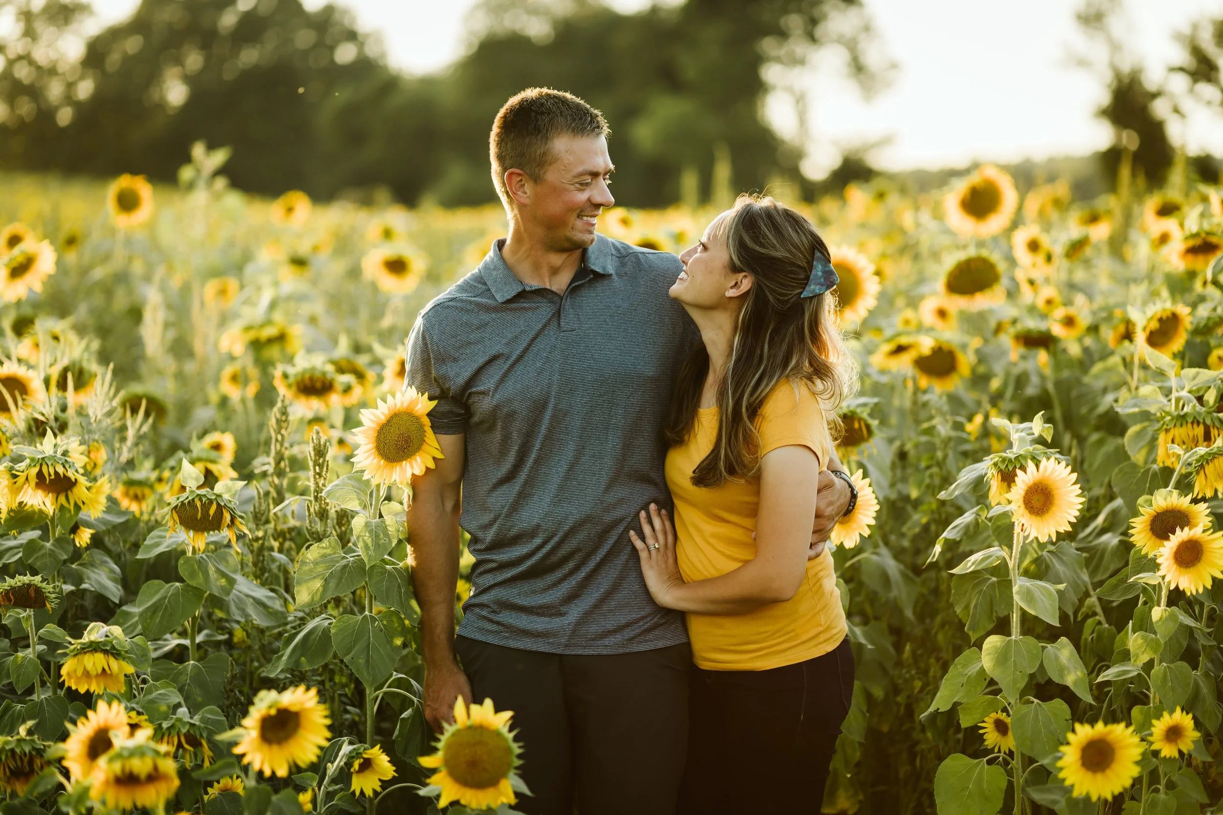 A couple standing in a sunflower field, smiling and looking at each other at sunset. Couple photography. Michigan.
