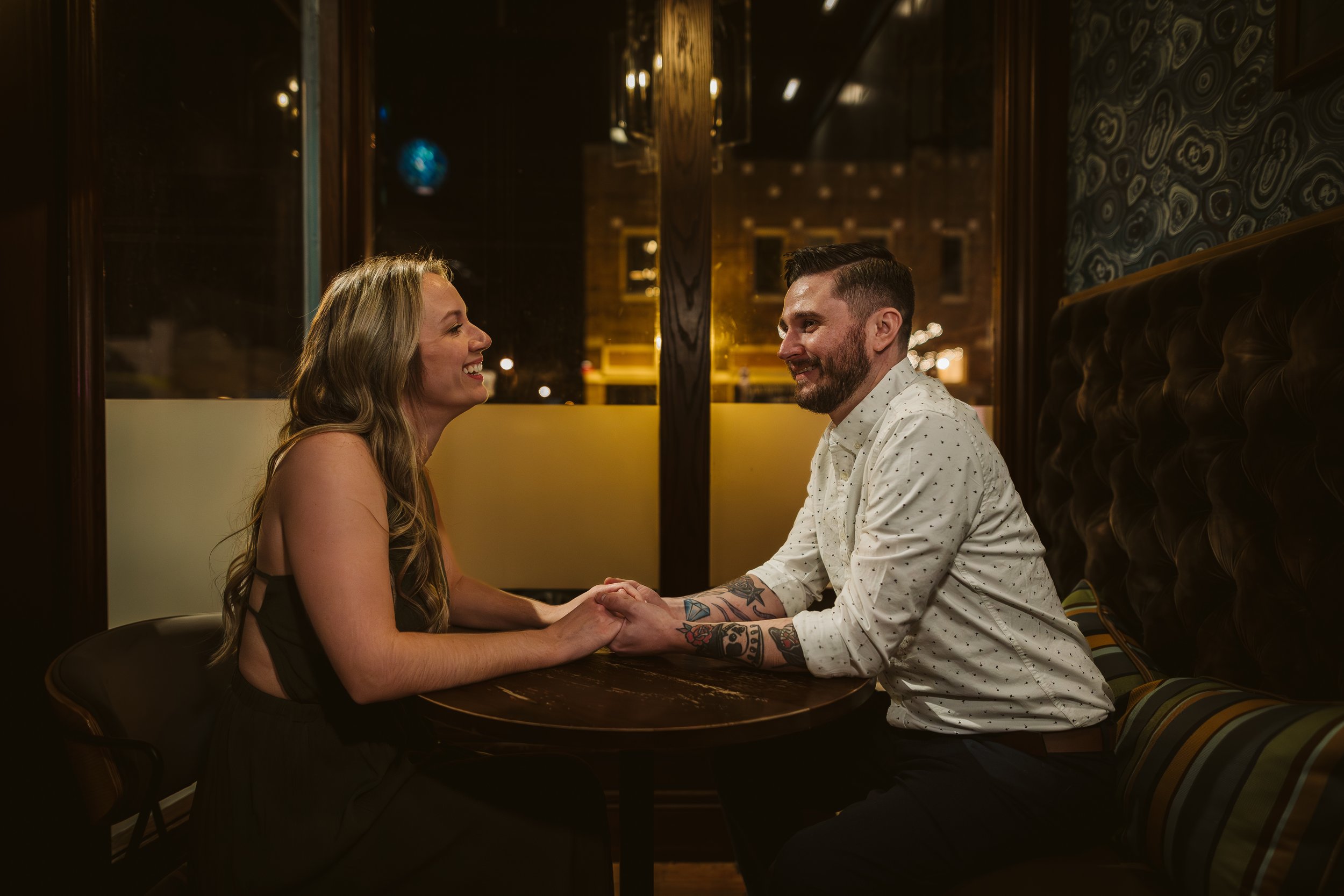 A couple sitting at a table in a dimly lit restaurant, holding hands and smiling at each other. They are enjoying a romantic moment with city lights visible through the window behind them. Engagement photoshoot.
