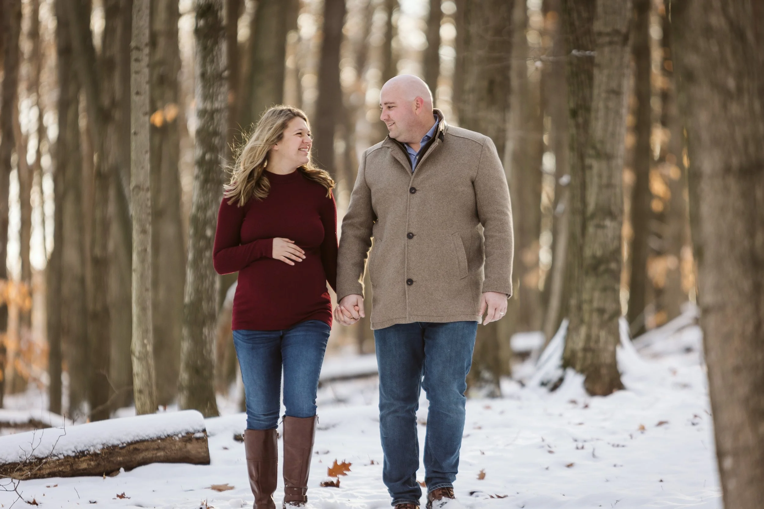 A man and woman holding hands and walking through a snowy forest, smiling at each other. Maternity photography. Cutlerville, Michigan.