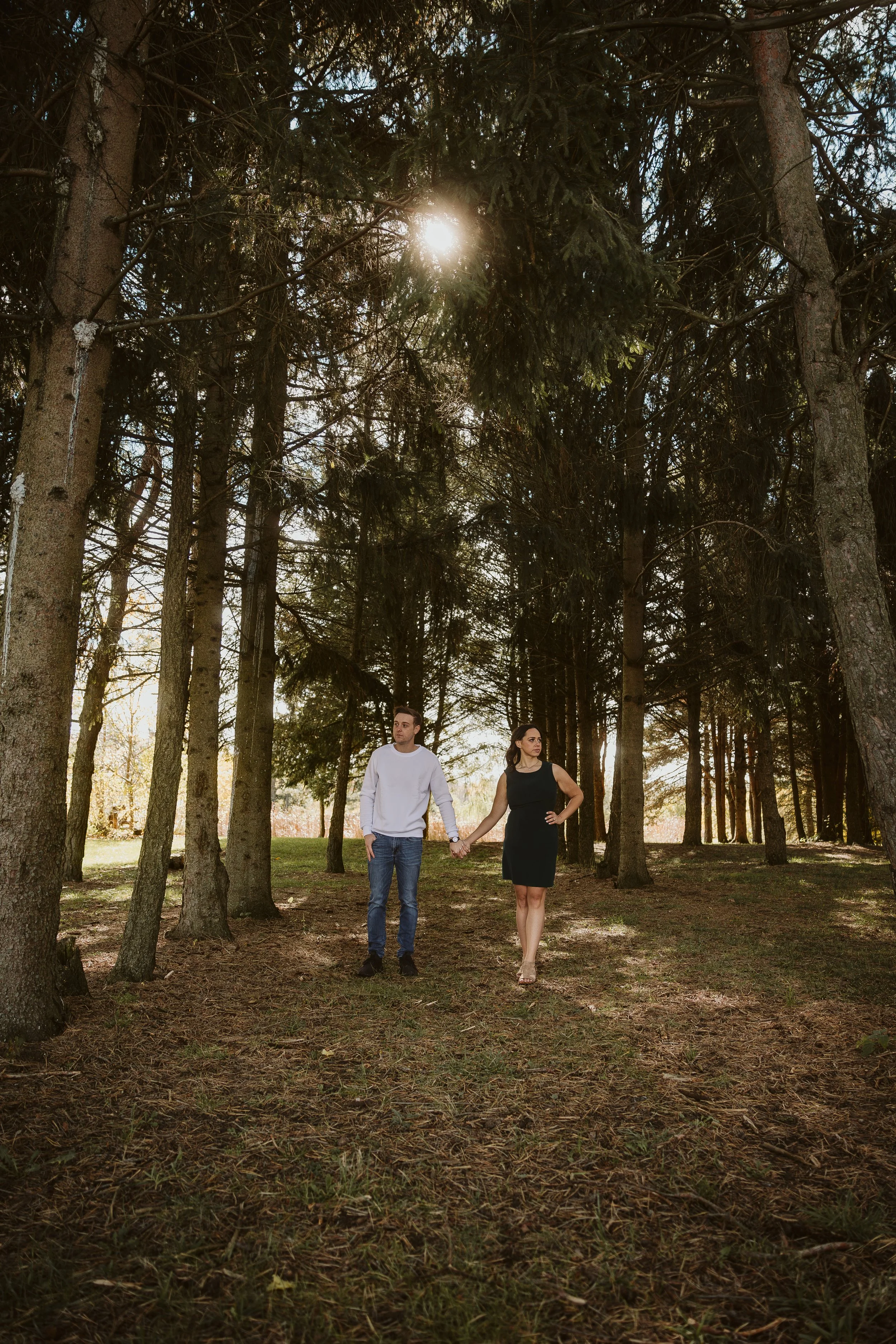 A couple walking hand in hand through a forested area during daytime, with the sun shining through the trees. Engagement photoshoot.