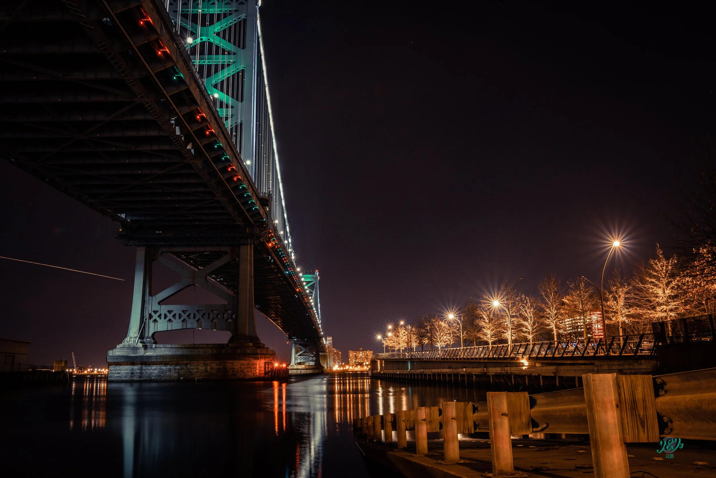 Night shot of a large suspension bridge over water, with colorful lights illuminating its cables, and a row of leafless trees with streetlights along a walkway to the right. Benjamin Franklin Bridge, Philadelphia, Pennsylvania. Landscape photography