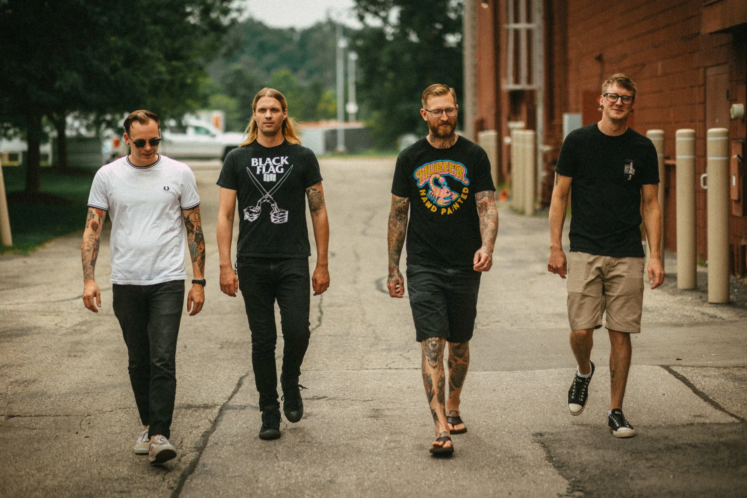 Four young men walking on a paved street near a brick building with trees in the background. Band photography. Grandville, Michigan.
