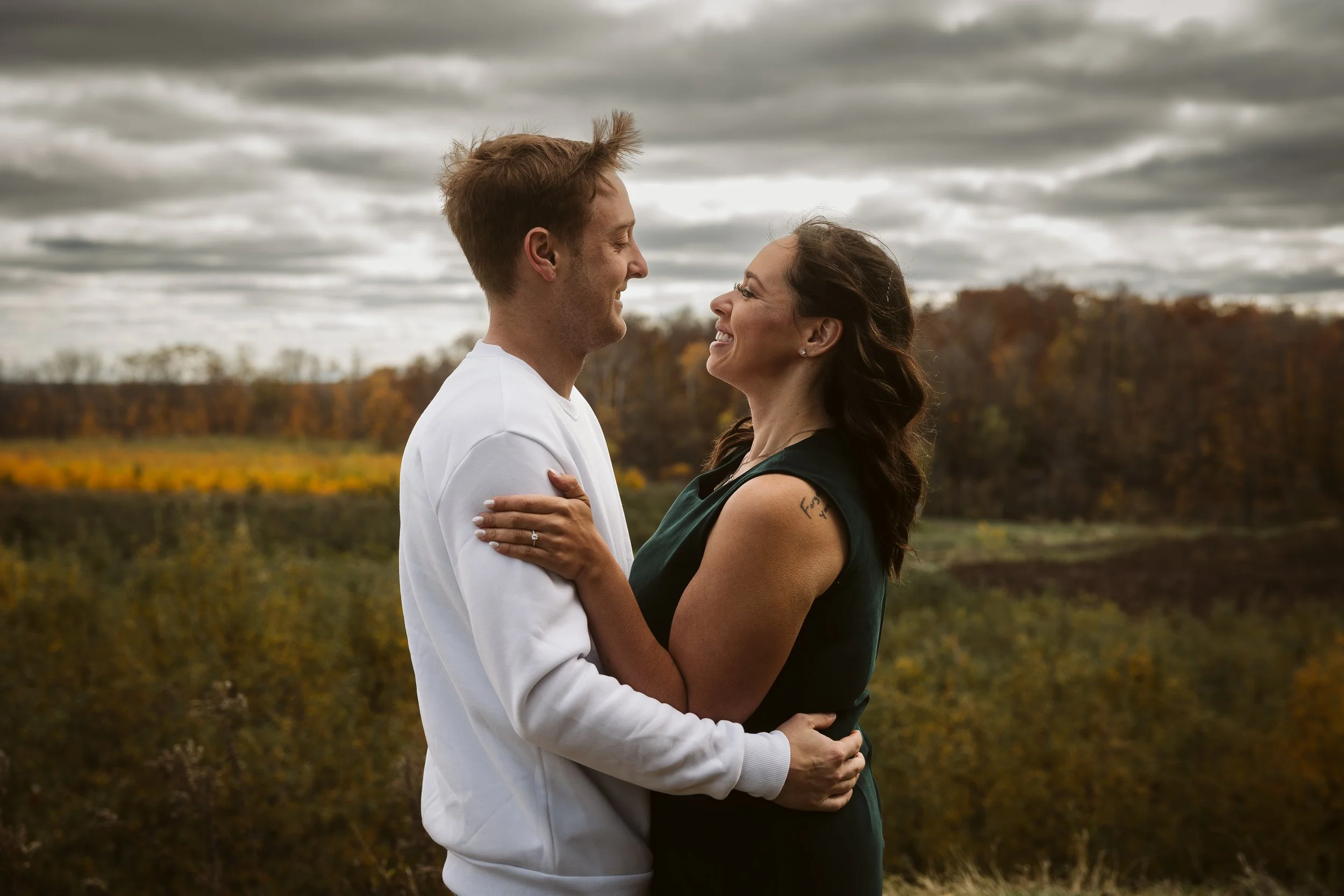 A smiling couple facing each other outdoors in a field with autumn trees in the background, holding each other. Engagement photoshoot.
