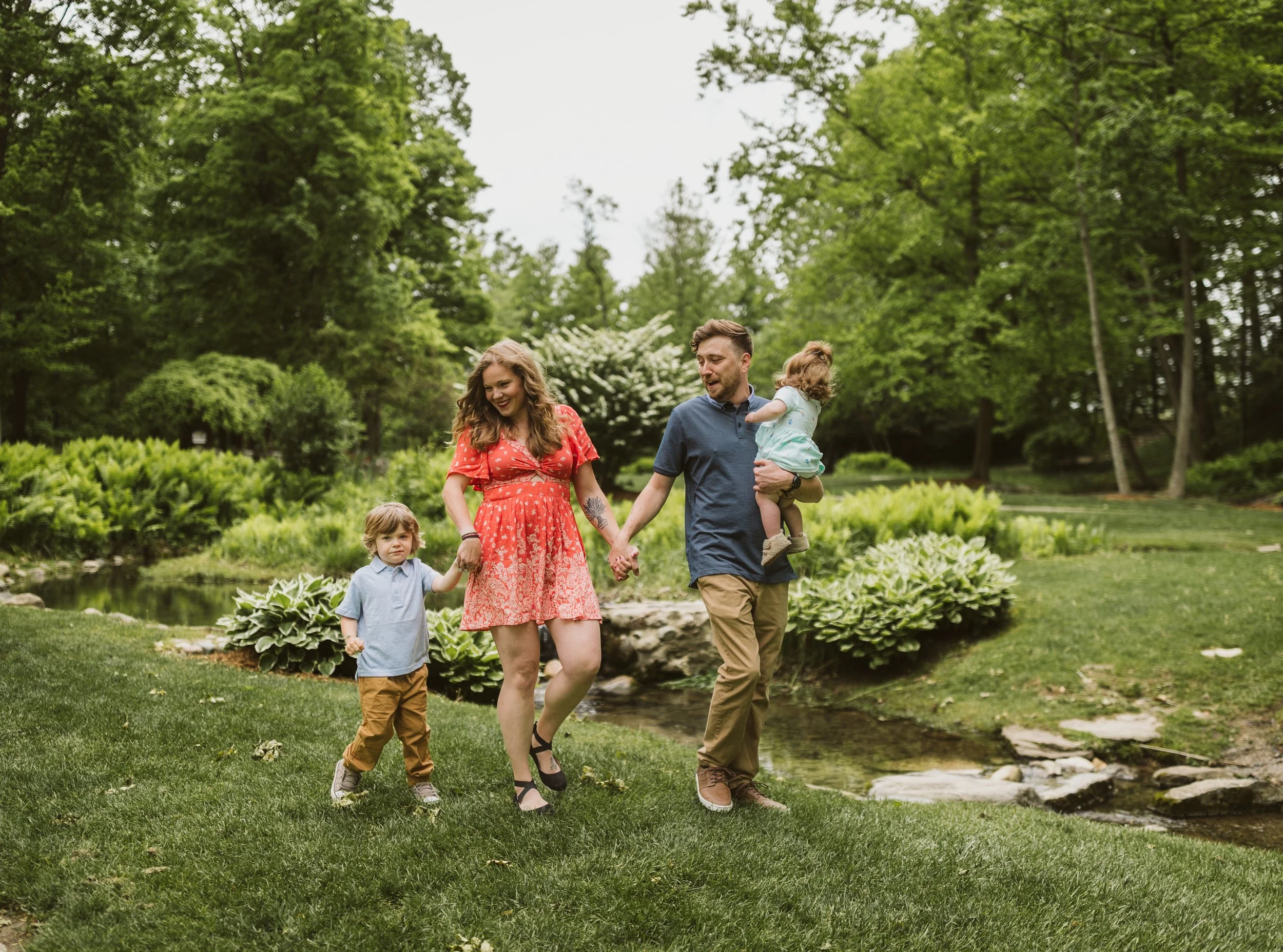 A family of four walking on a grassy area near a small stream in a park, holding hands and smiling, surrounded by green trees and bushes on a cloudy day. Family photography. Grandville, Michigan.
