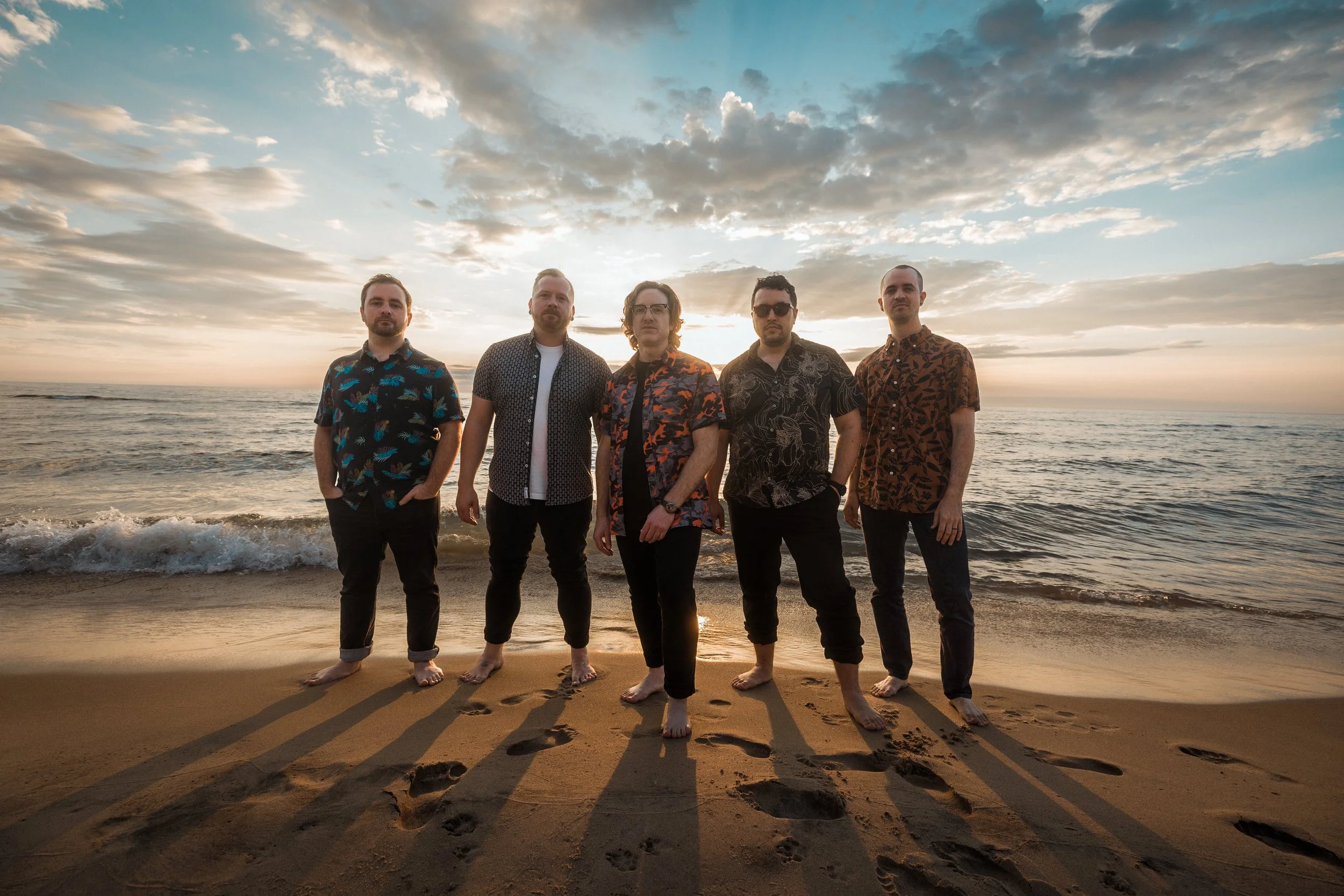 Five men standing barefoot on a sandy beach at sunset, with ocean waves behind them and clouds in the sky. Lake Michigan. Band photography.
