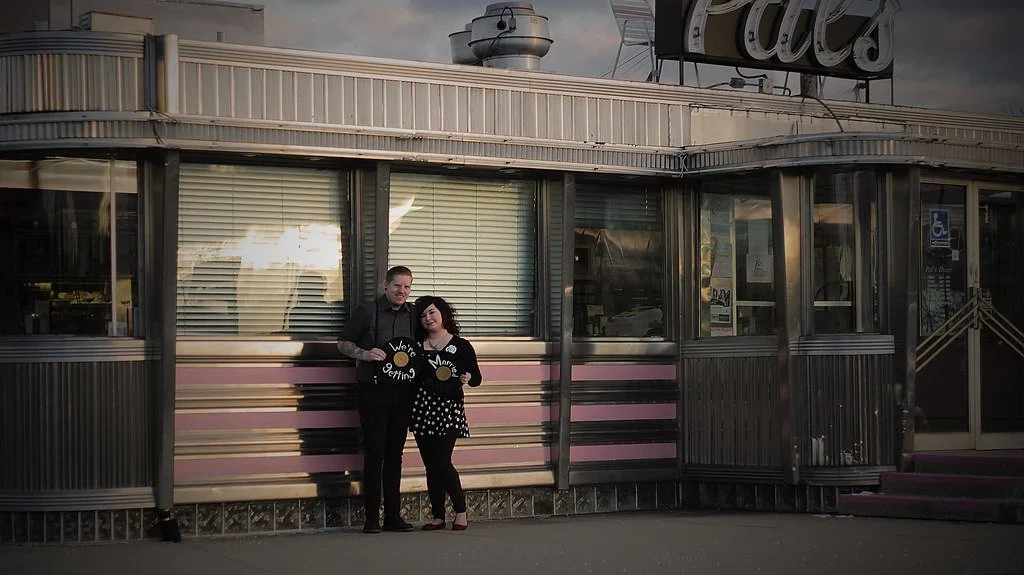 Two people standing in front of a vintage diner with a pink and black striped exterior. Engagement photoshoot.
