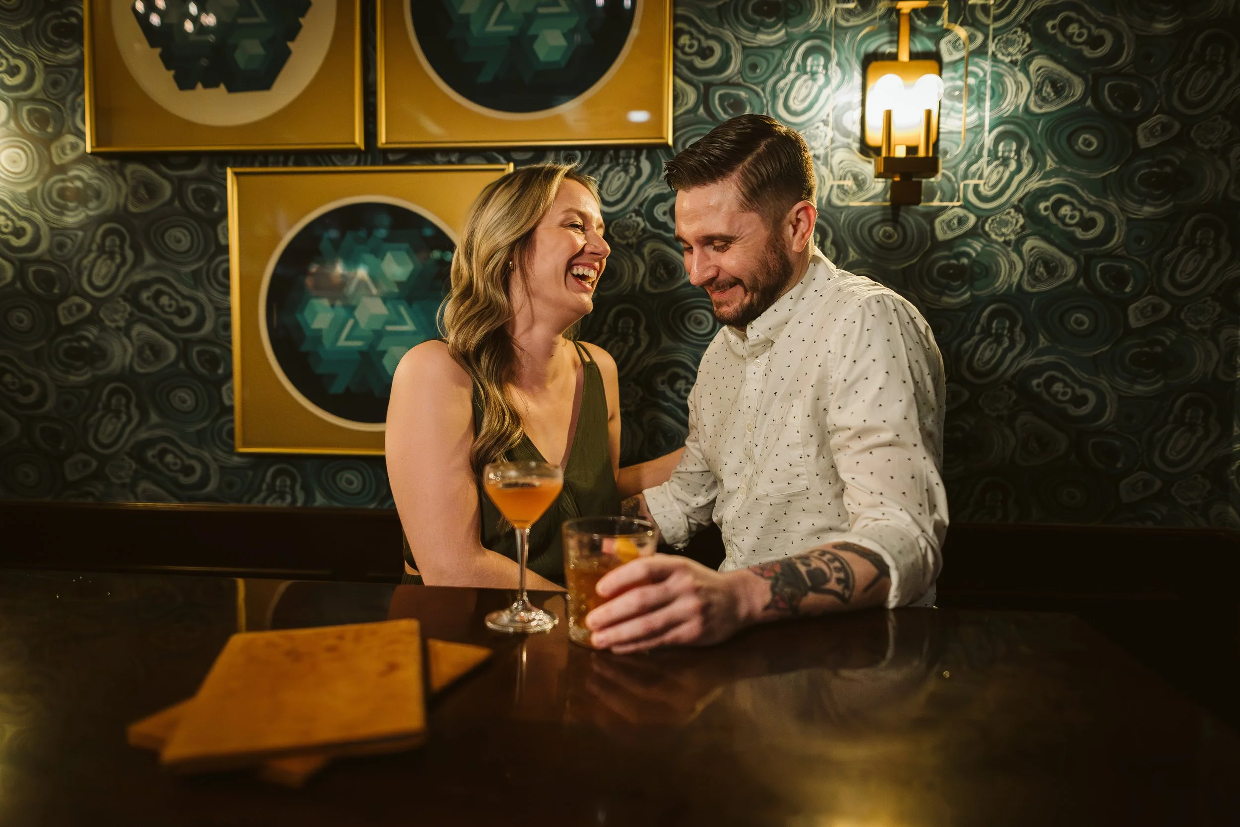 A smiling woman and man enjoying drinks together at a bar, with colorful artwork on the wall behind them. Engagement photoshoot.