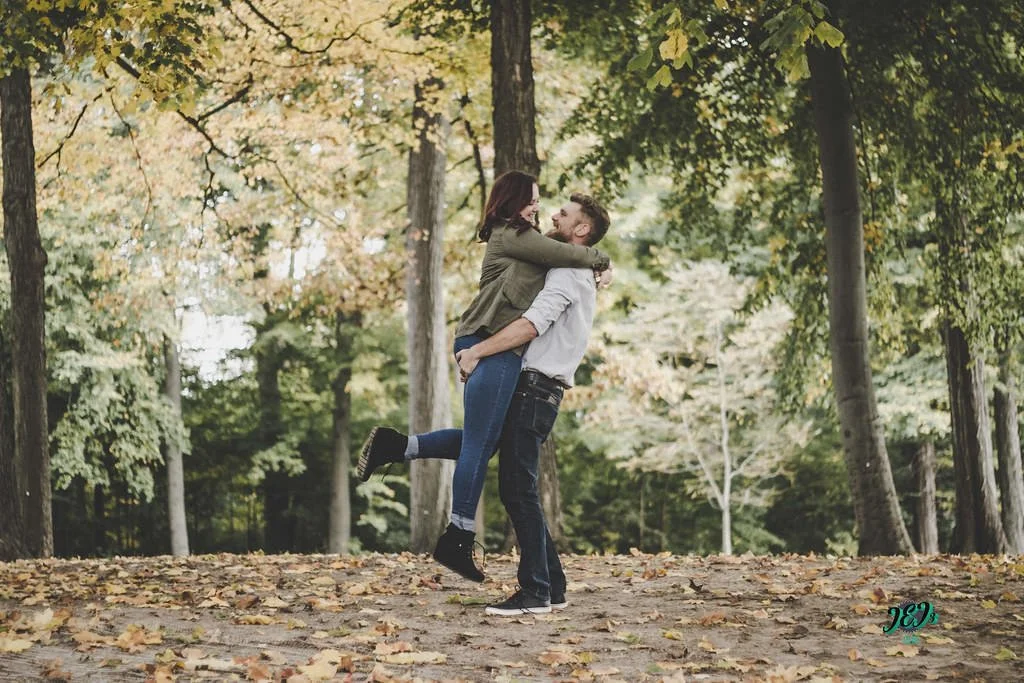 A couple happily embracing in a forest with autumn leaves on the ground and trees with colorful fall foliage. Engagement photoshoot.