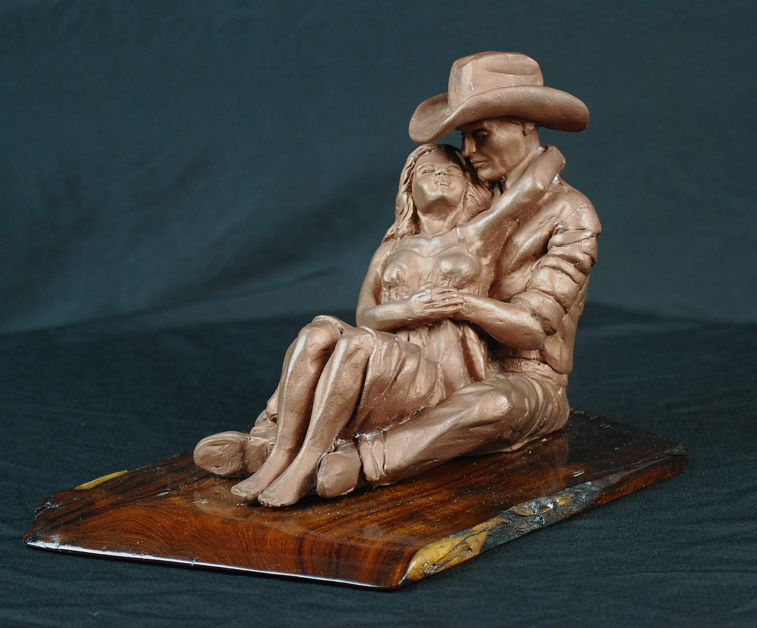 Cowboy Couple sculpture by Sharon Cruson, seated Western couple embracing on a natural wood base against a dark background