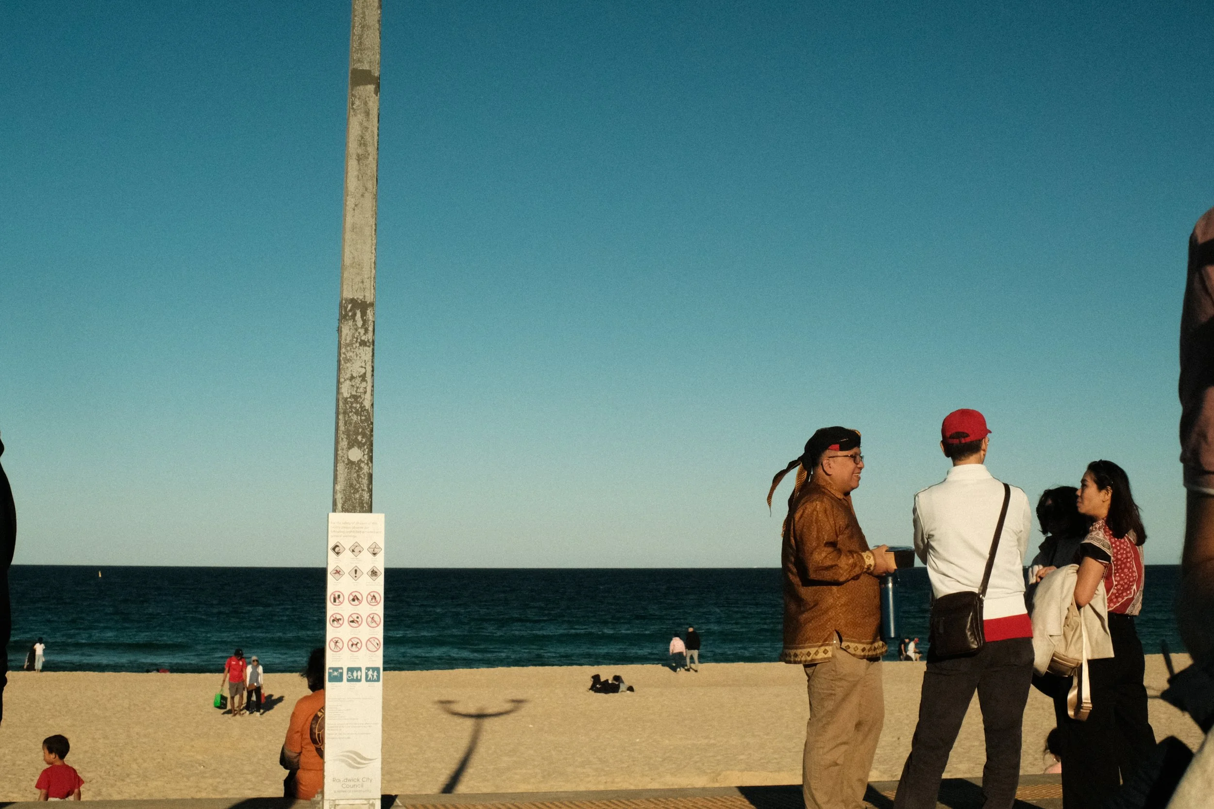 [After The Flag Comes Down by Fadlan Hamizan Ekantoro] -  You might look at this picture and think this is a normal photograph of a sunny afternoon on Maroubra Beach. But look closer and you’ll see bits and pieces of home. A blangkon. Batik. Red and 