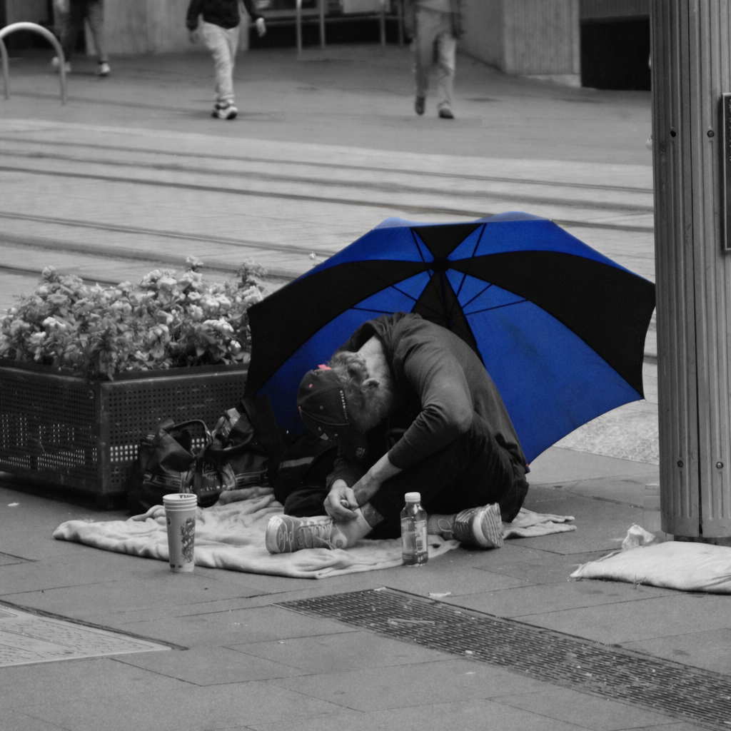 [The Blues by Finlay McCreath] - For this image, I chose a black and white street scene with the umbrella kept in bright blue to create a strong contrast. The person sitting on the pavement represents homelessness and the struggles that can exist wit