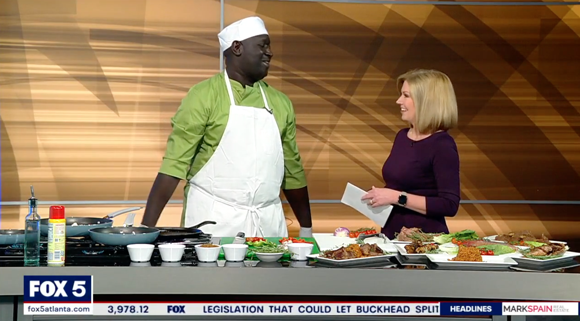 A TV show segment on FOX 5 featuring a male chef and a female host in a kitchen studio. The chef is wearing a green shirt, white apron, and chef hat, while the host is dressed in a purple top. They are standing behind a counter with various dishes, ingredients, and cooking utensils. The background has a wooden pattern, and a news ticker is visible at the bottom of the screen.