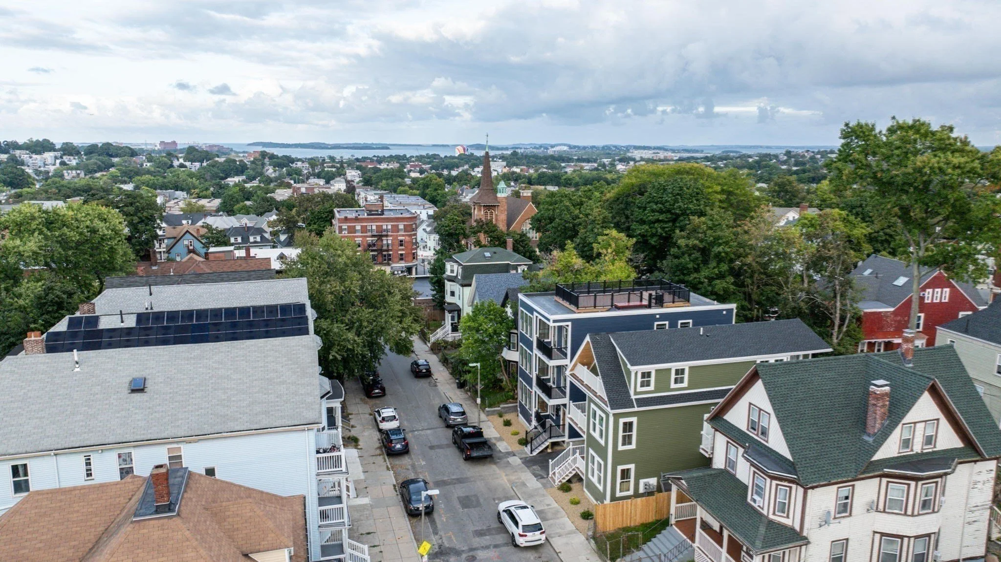 Aerial view of a neighborhood with residential houses, trees, and a church with a tall steeple, under a cloudy sky.