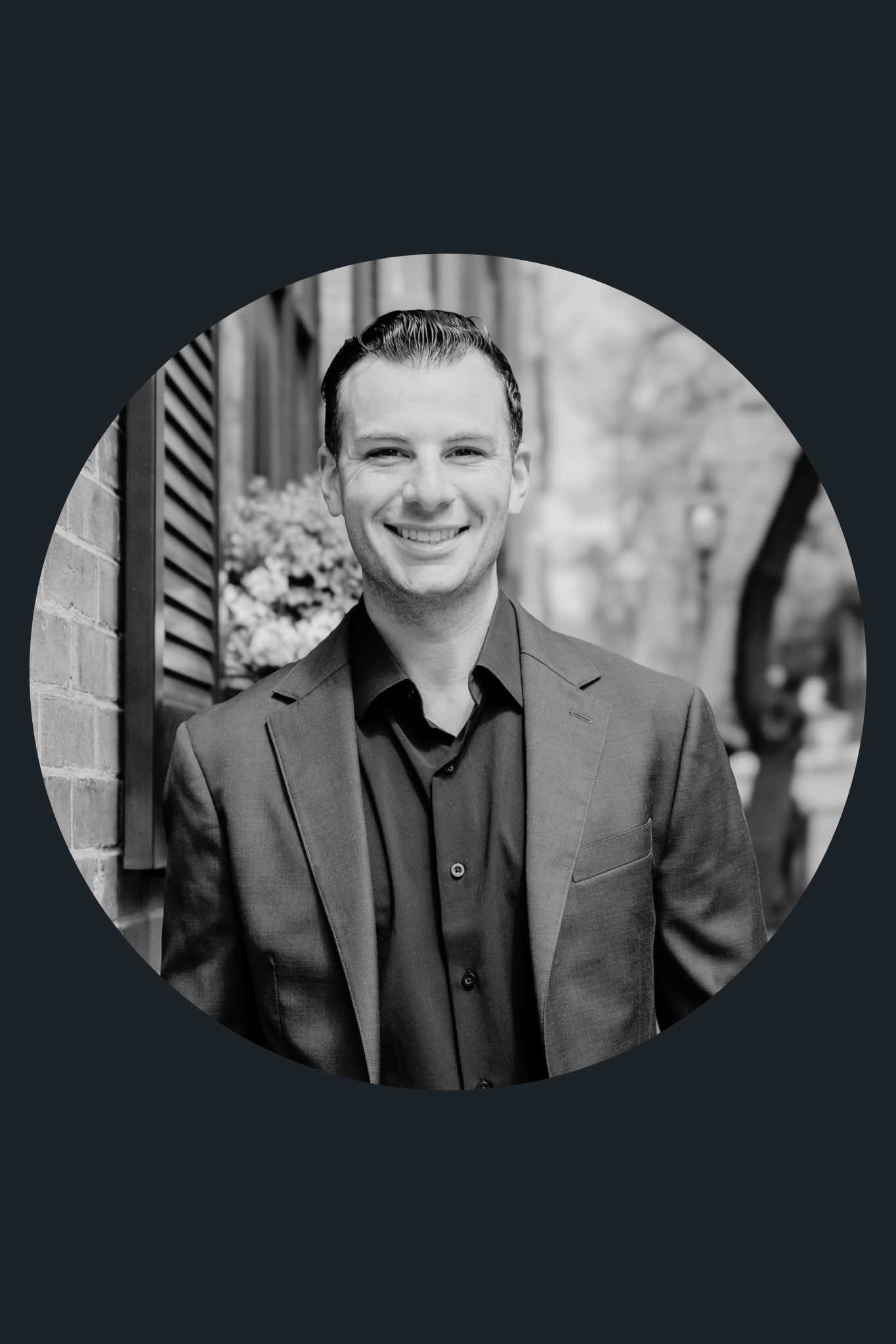 Black and white portrait of a smiling man in a suit, standing outdoors near a brick wall and wooden window frame, with trees and a street in the background.