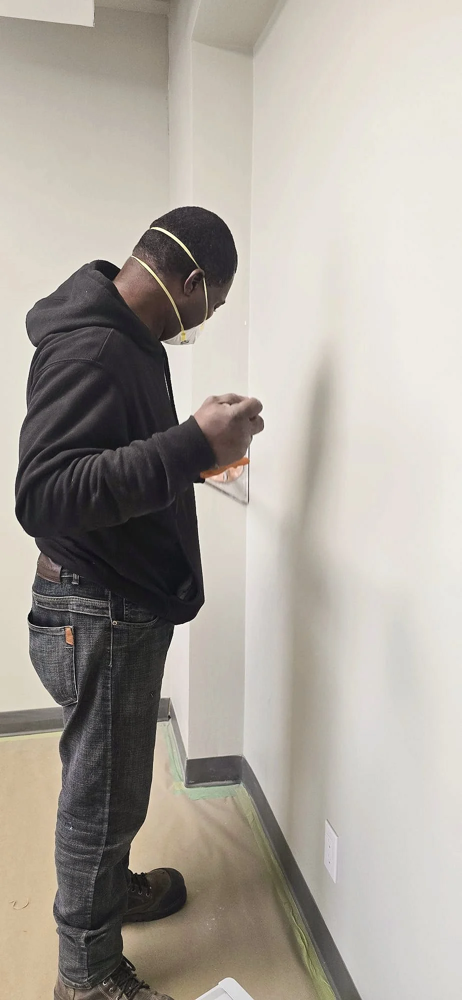 A Red Oak Painting 101 Program participant standing indoors wearing a black hoodie, dark jeans, brown boots, and a face mask, painting a corner wall.