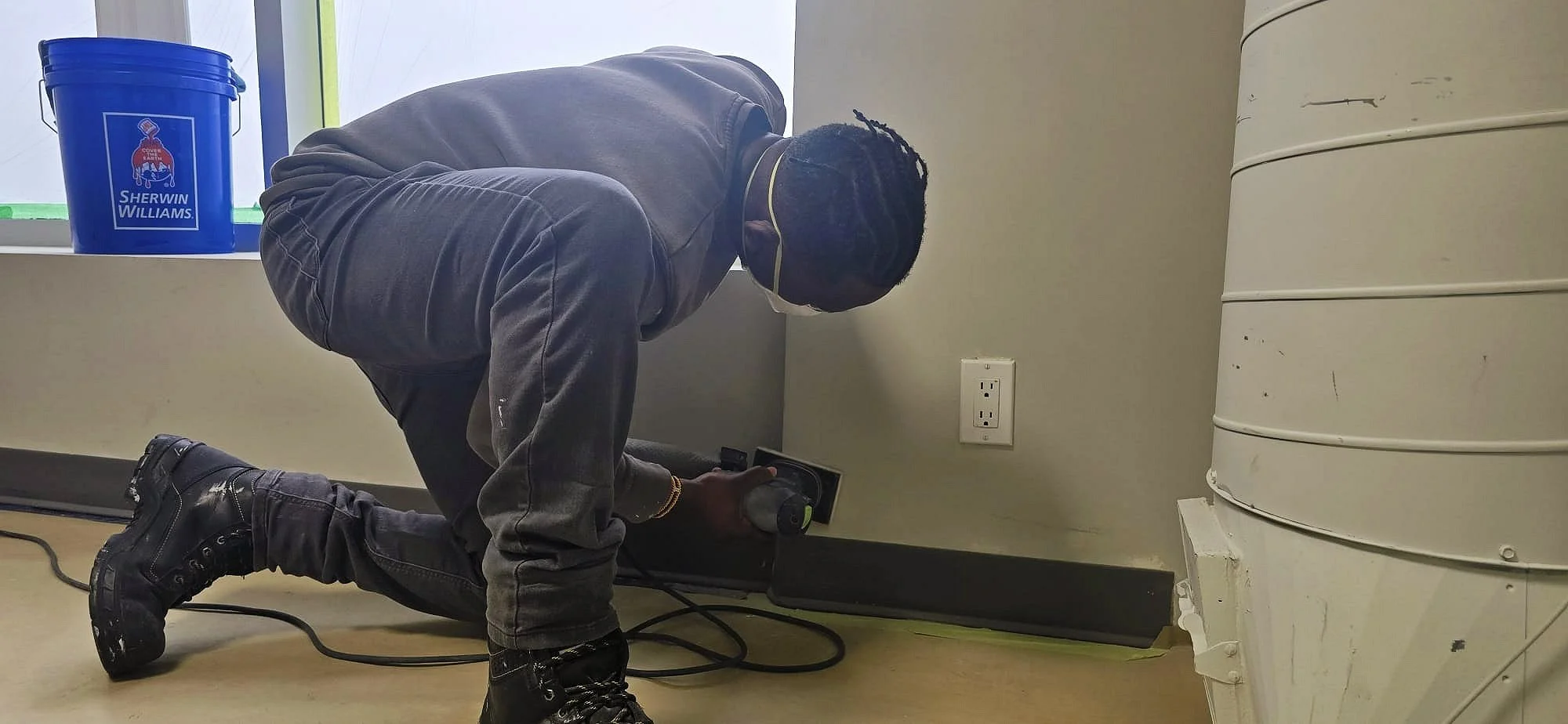 A man kneeling on the floor fixing or inspecting a power outlet with a power drill, wearing a face mask, a dark jacket, jeans, and black boots. There is a blue Sherwin Williams paint bucket on the window sill nearby.