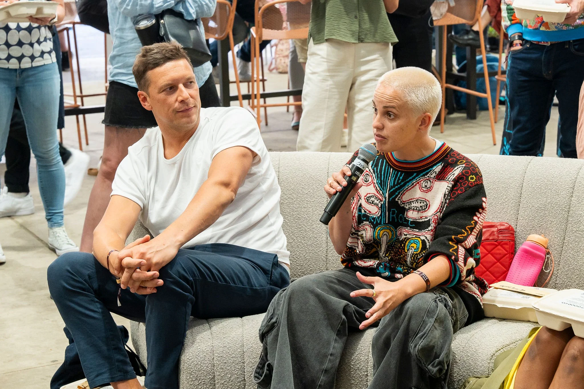 A person with short hair, wearing a colorful patterned sweater, is speaking into a microphone while seated on a couch. Another person in a white t-shirt is sitting beside them. Several people are standing in the background, including one holding a takeout container and another holding a reusable cup.