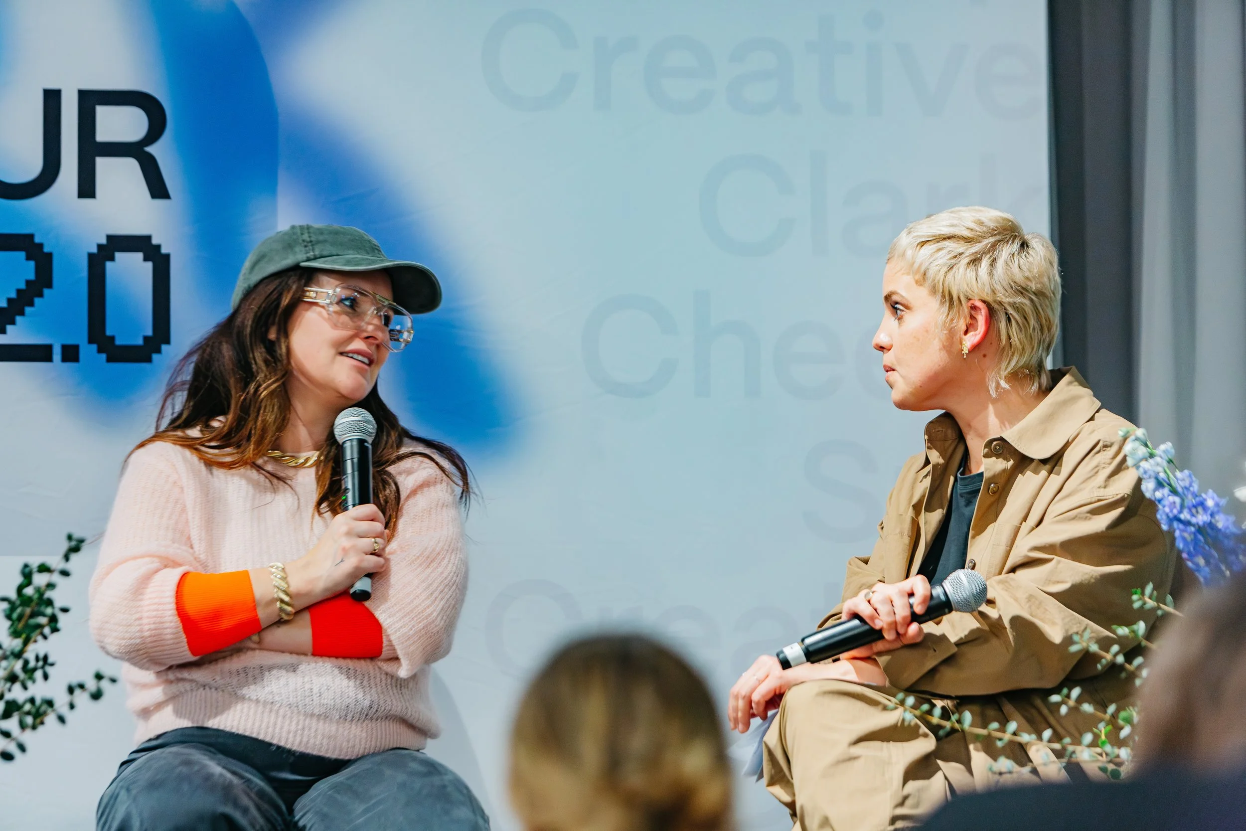 Two women sitting and talking on stage with microphones during a discussion event. One wears a pink sweater and cap, the other in a tan outfit. There is a banner in the background with some text.