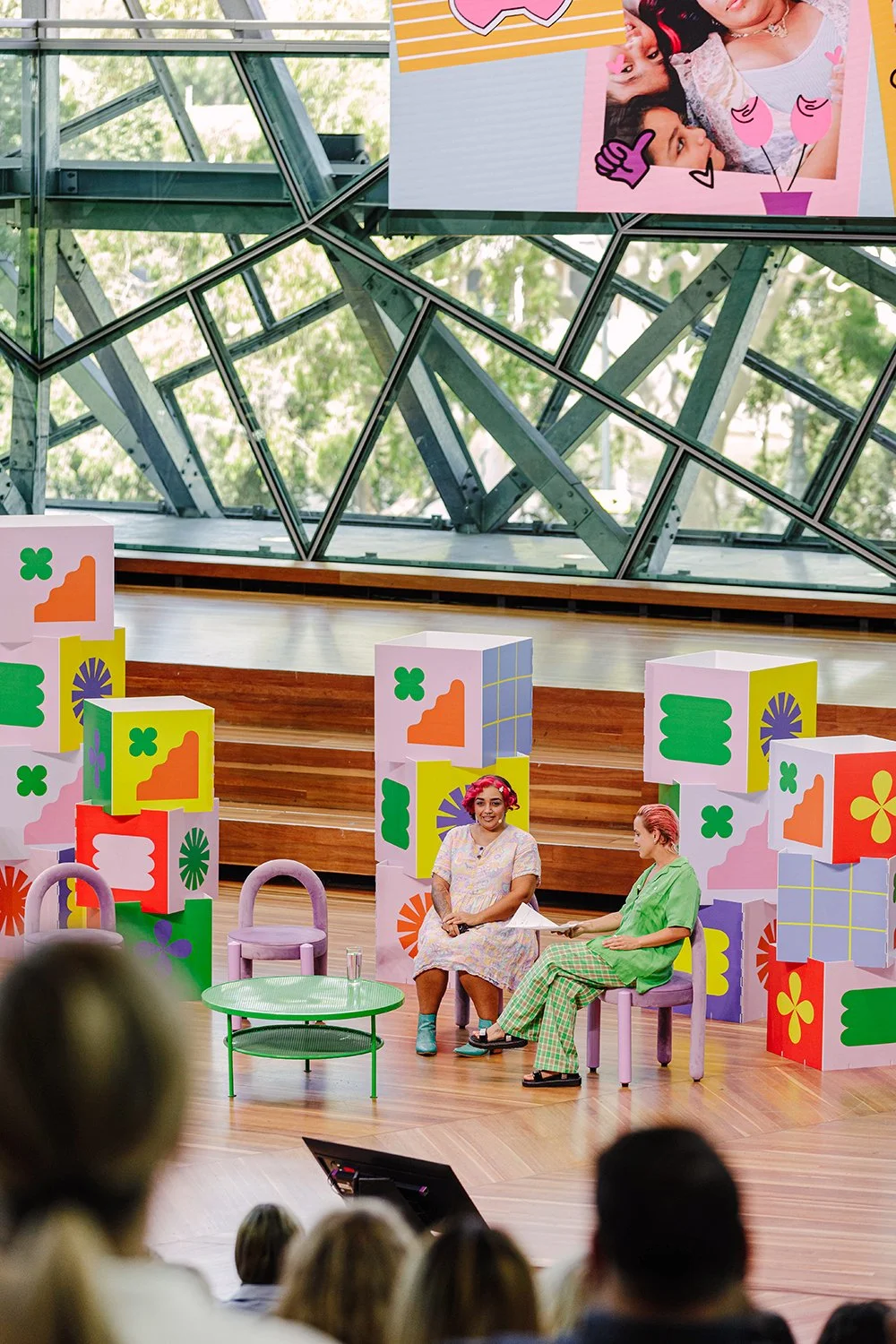Two people sitting on stage in a colorful setting with geometric and floral decorations, speaking to an audience. A large screen above them displays a photo collage and graphic elements.