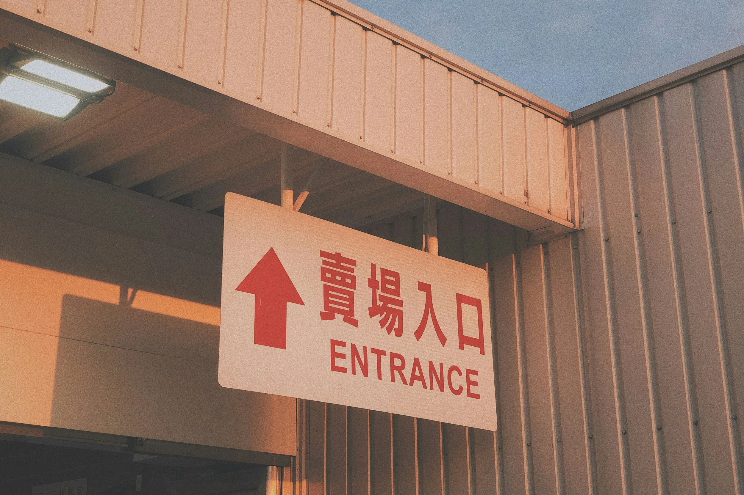 A building entrance sign with Chinese characters and the word 'ENTRANCE' in English, featuring a red arrow pointing upward, under a metal awning with evening lighting.