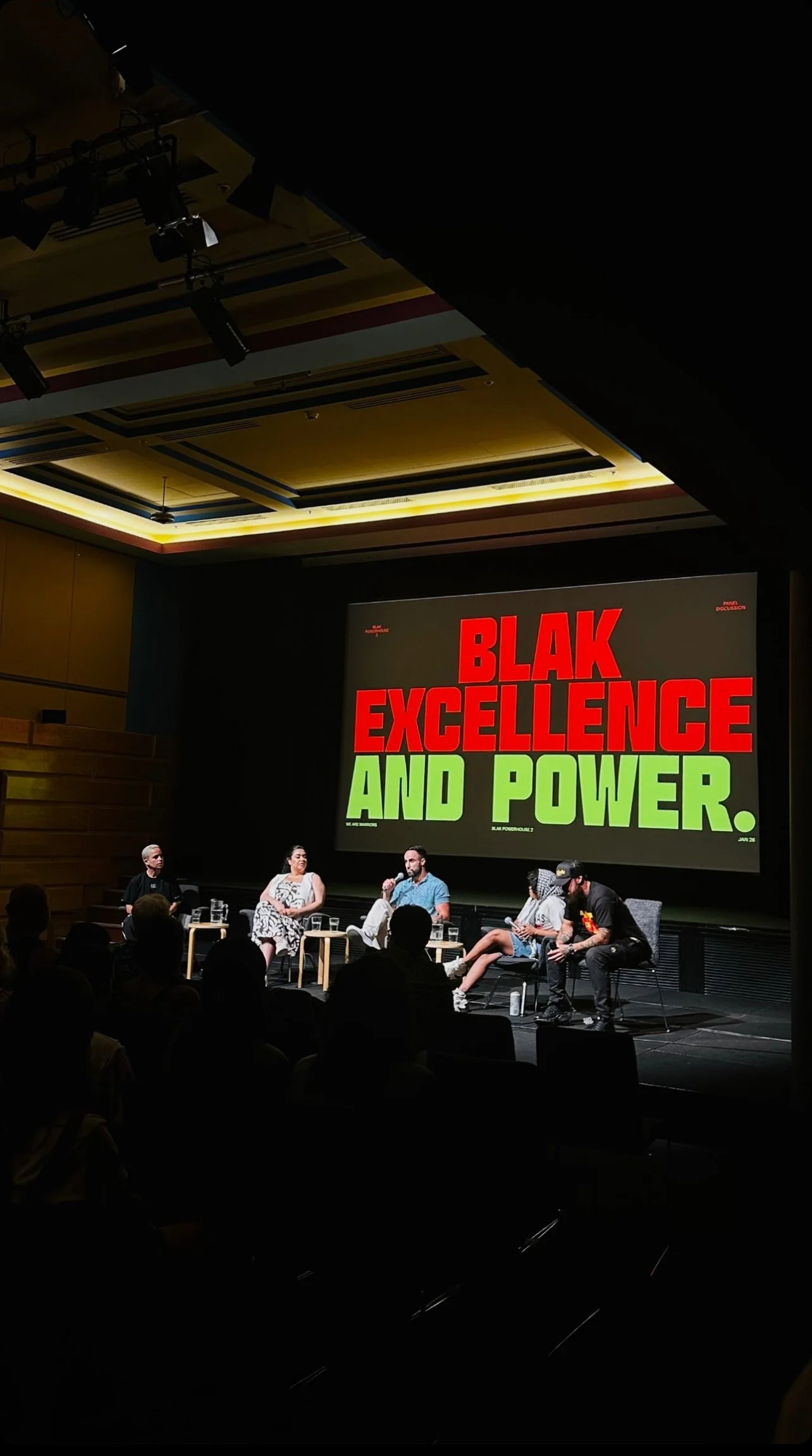 Panel discussion event titled 'Blak Excellence and Power' with five speakers seated on stage, audience in foreground. Large screen behind displays event title in bold red and green text.