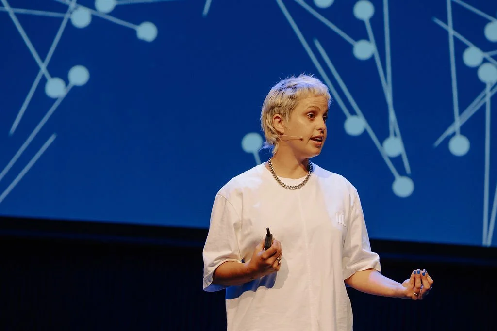 Person giving a presentation on stage with a blue background showing a network-like pattern, holding a microphone and gesturing with hand.