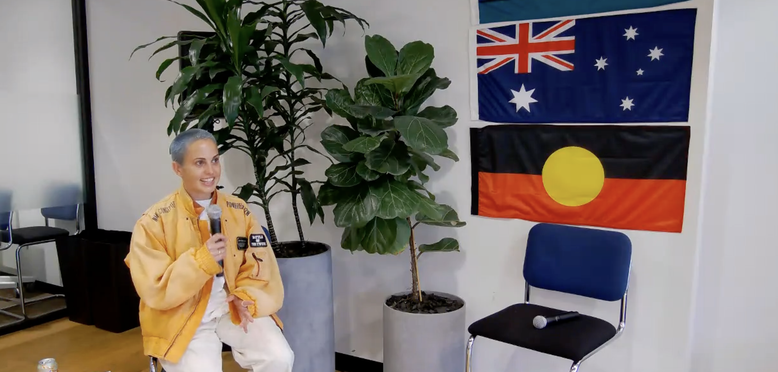 Person in yellow jacket holding a microphone, seated near two potted plants, in front of an Australian and an Aboriginal flag, with an empty chair and a second microphone beside them.